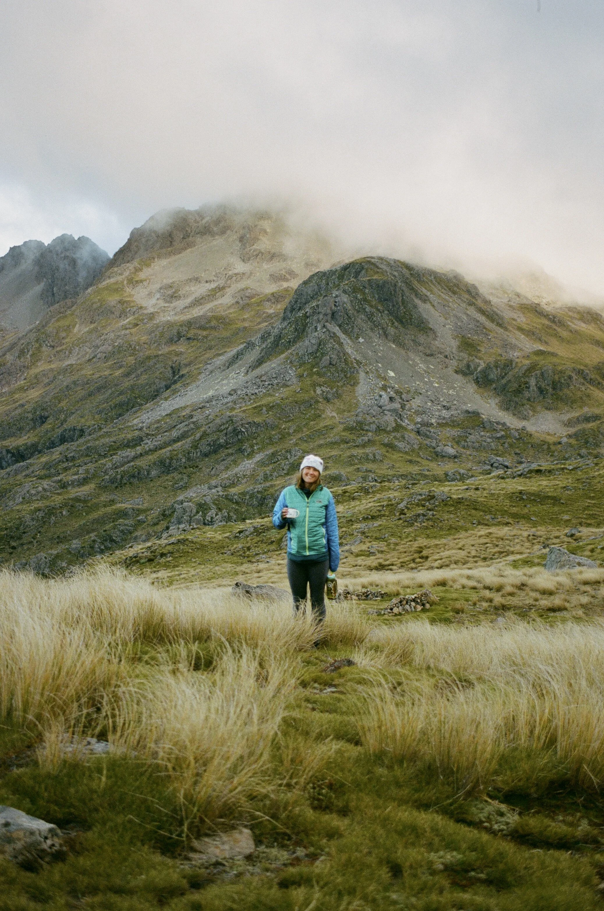 A woman stands in a grassy field with mountains partly covered in clouds in the background, holding a coffee mug and wearing outdoor hiking clothing.