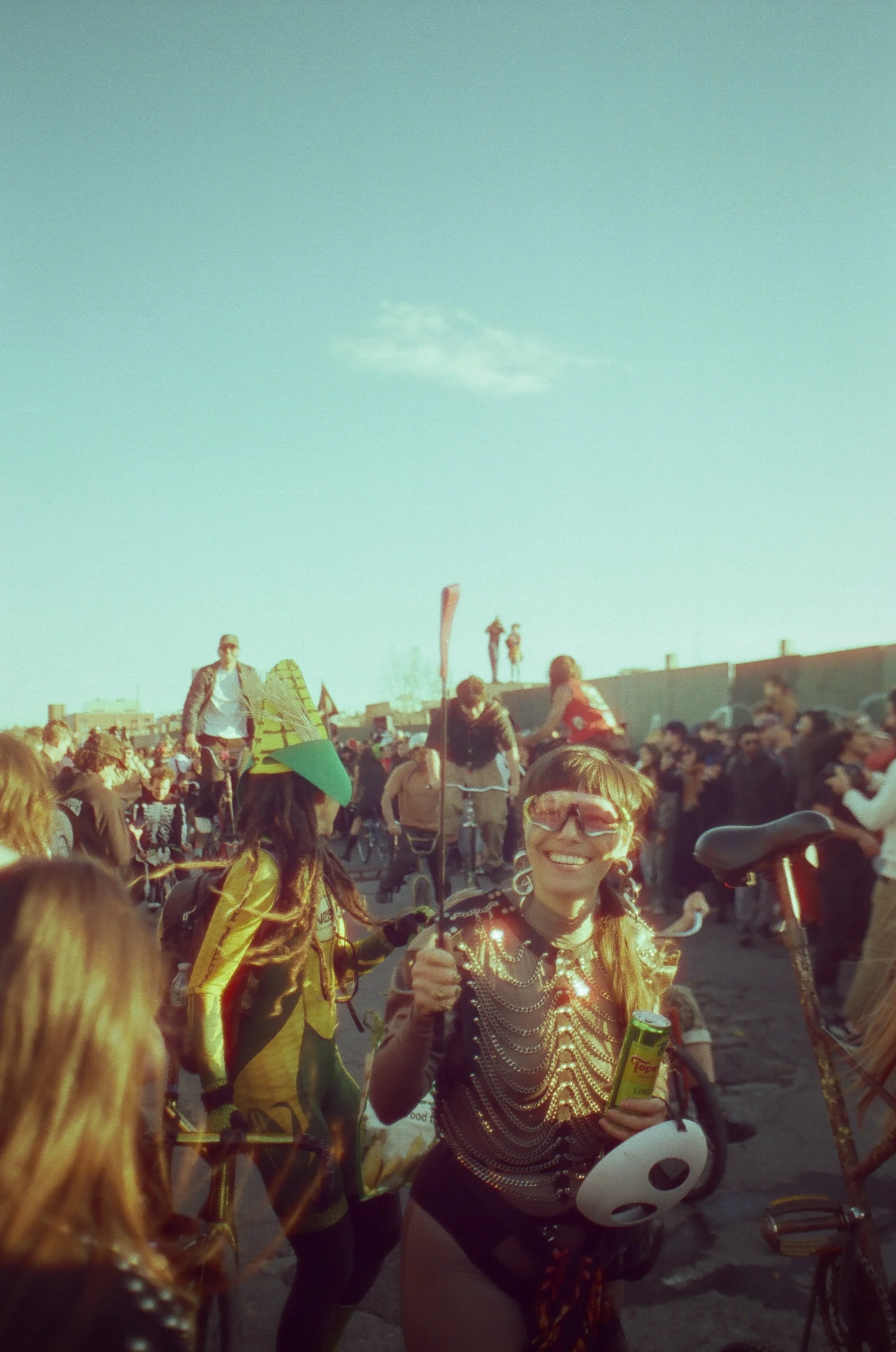 A woman dressed in a skeleton costume smiling, holding a small cake and a balloon, at an outdoor event with many people.