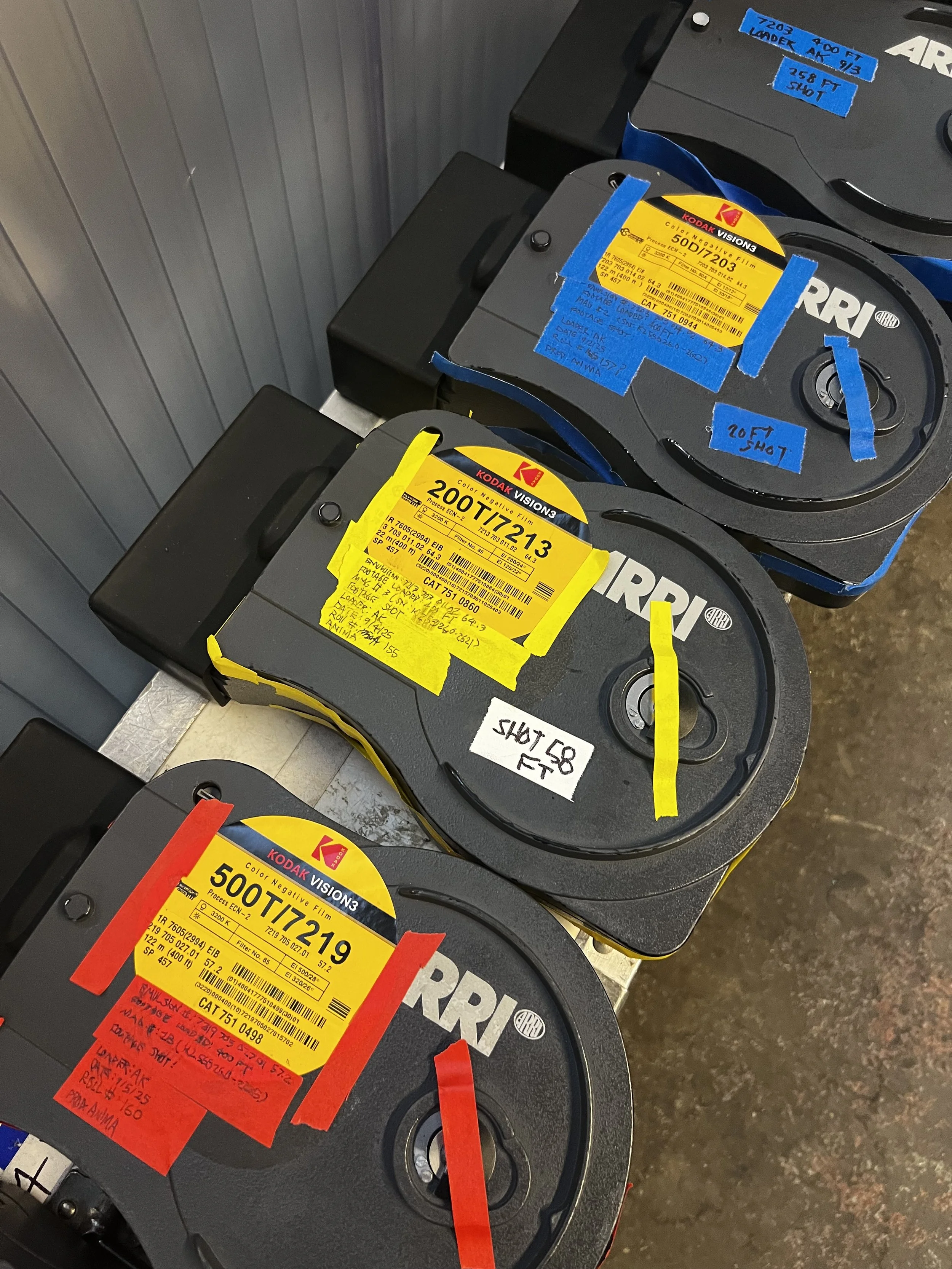 Three large automotive parts labeled with yellow, blue, and red stickers, placed on a metal surface next to a gray wall. The stickers contain part numbers and other information.