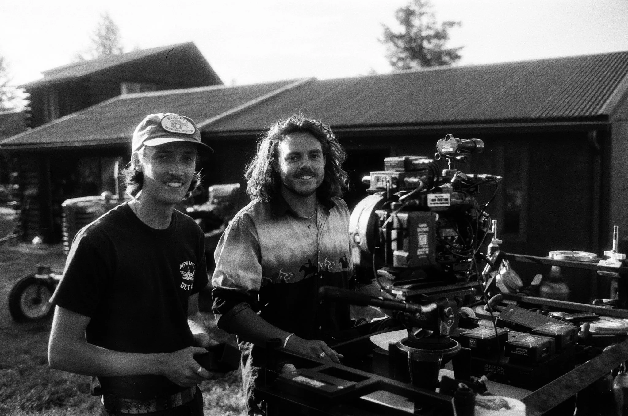 Two men on a film set outdoors, one holding a small device, with a camera and other equipment in front of them, a house and trees in the background.