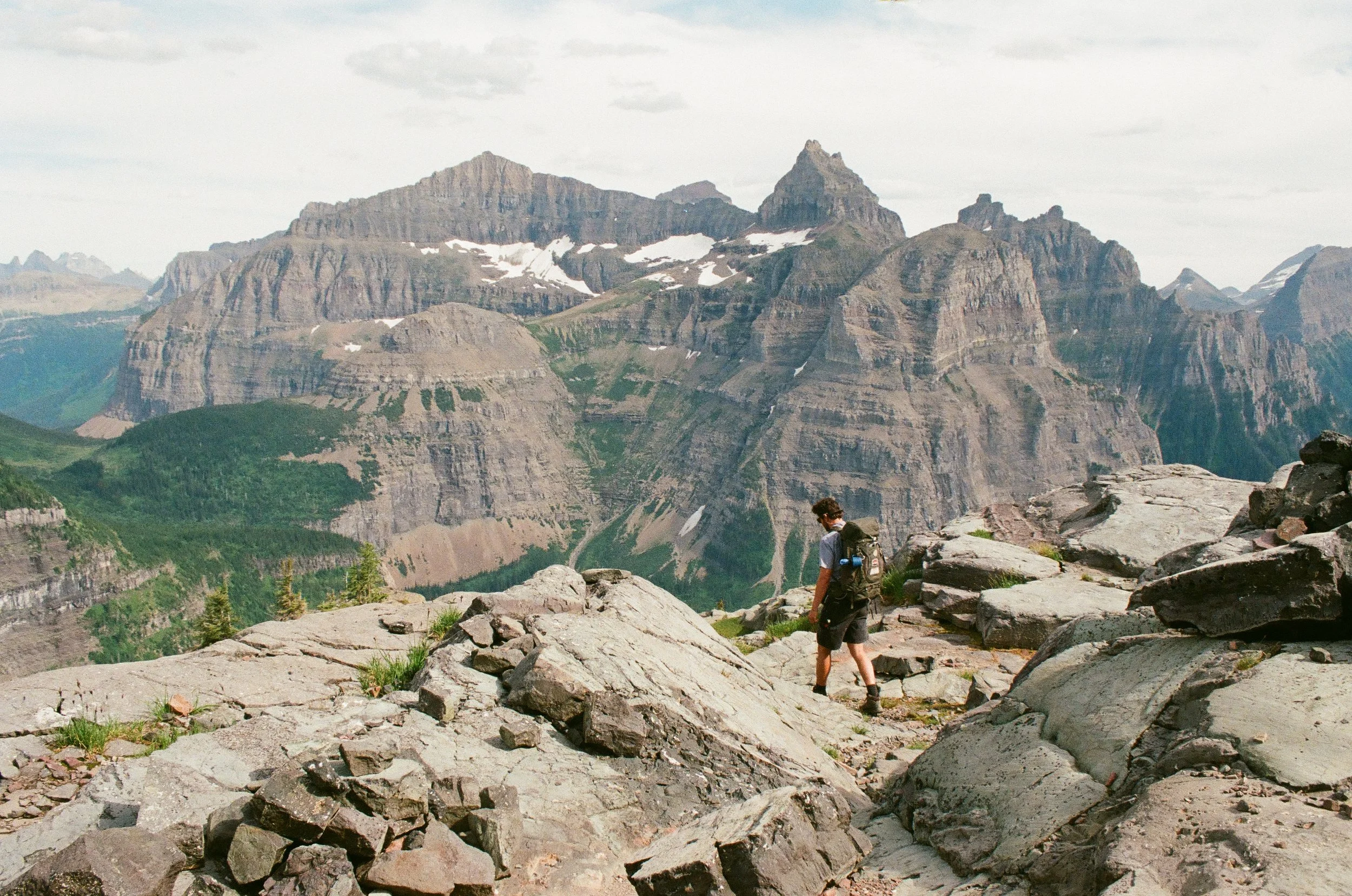 A person with a backpack hiking on a rocky trail in a mountainous landscape with tall peaks and patches of snow.