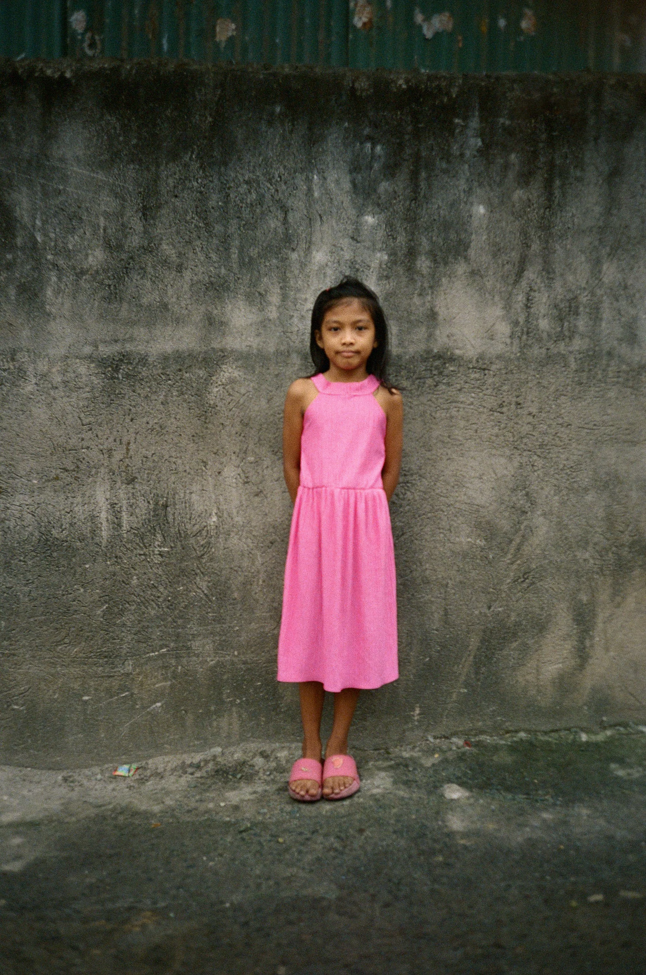 A young girl wearing a pink sleeveless dress and pink sandals stands against a weathered gray concrete wall.