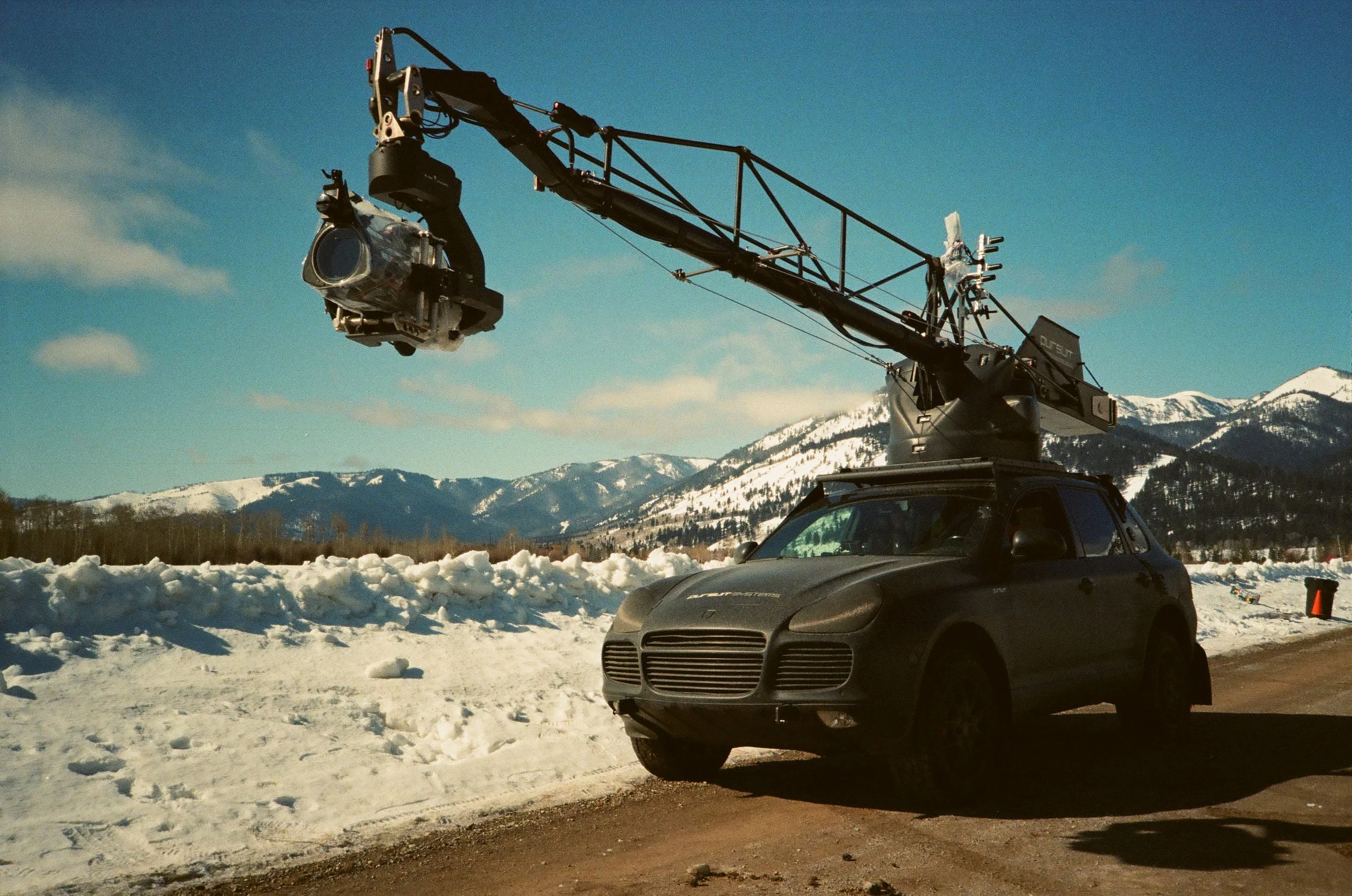 A professional camera mounted on a crane attached to a black car, set against a snowy mountain landscape with blue skies.