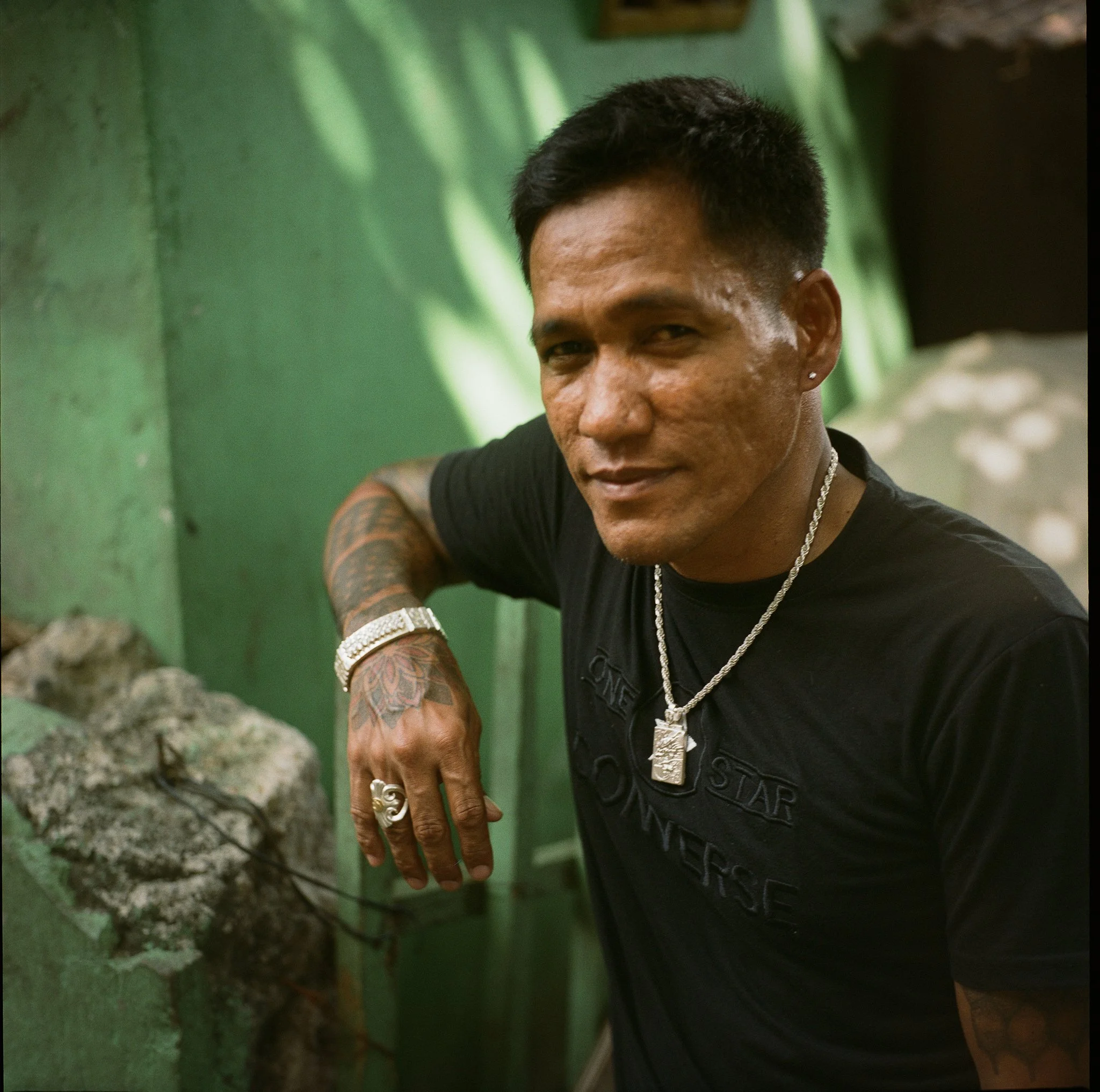 A man with tattoos, wearing a black t-shirt and silver jewelry, leans on a green wall outdoors.