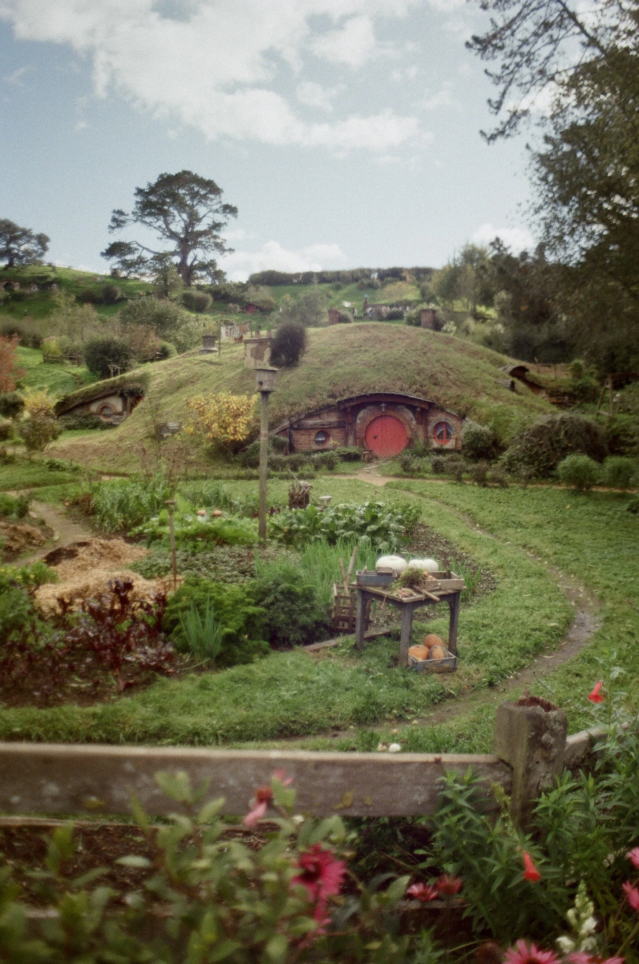 Hobbit-like house built into a grassy hillside with a circular red door, surrounded by garden and pathways on a cloudy day.
