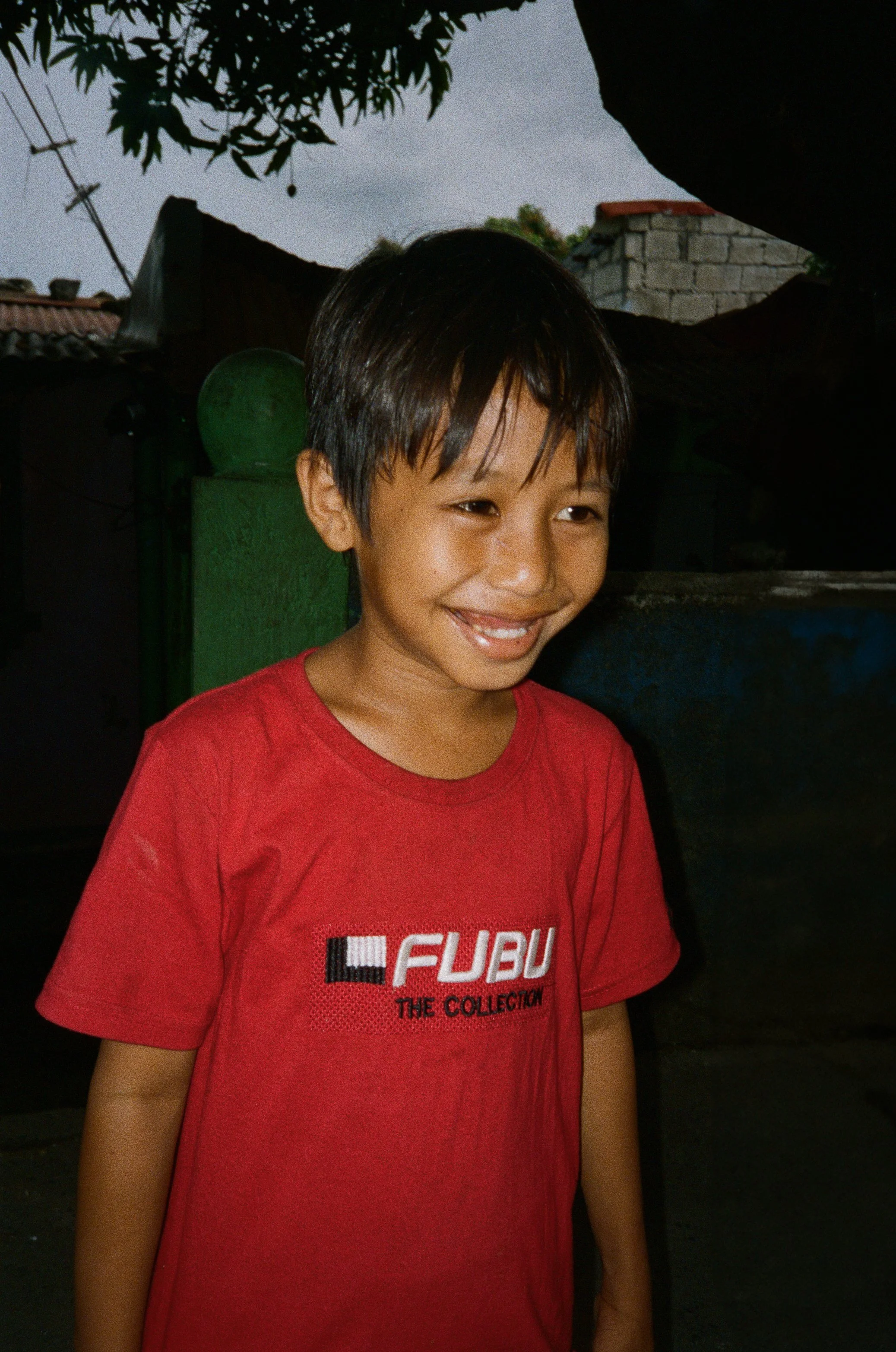 A smiling young boy with short black hair wearing a red T-shirt with the words "FUBU" and "The Collection" standing outdoors with a tree, a green gate, and a brick wall in the background.