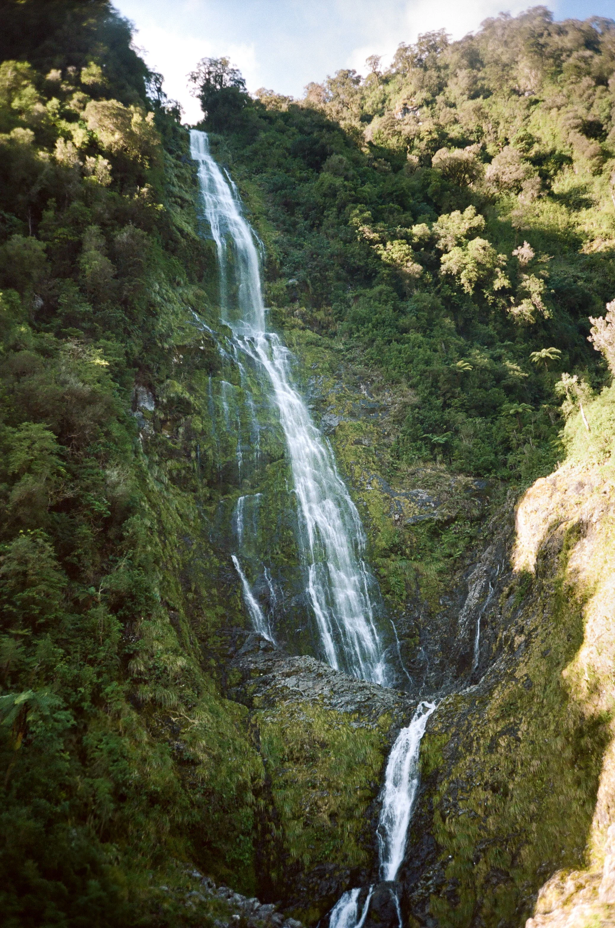 Tall waterfall cascading down a lush green mountainside surrounded by trees and vegetation.