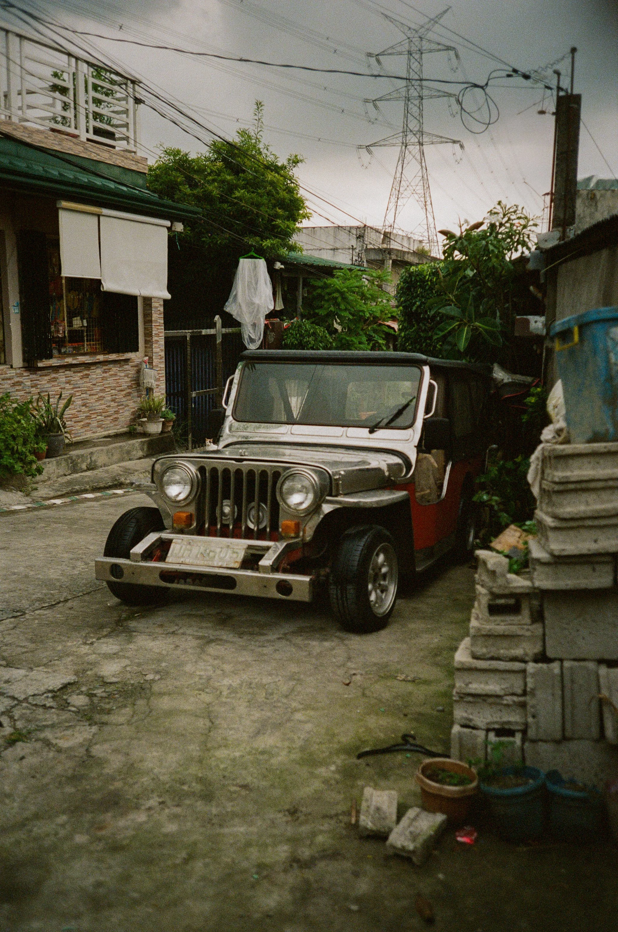 A vintage Jeep parked on a weathered concrete driveway in a residential area, surrounded by potted plants, a stone wall, and nearby buildings with laundry hanging outside. Overhead, power lines and a tall electrical tower are visible against the clou