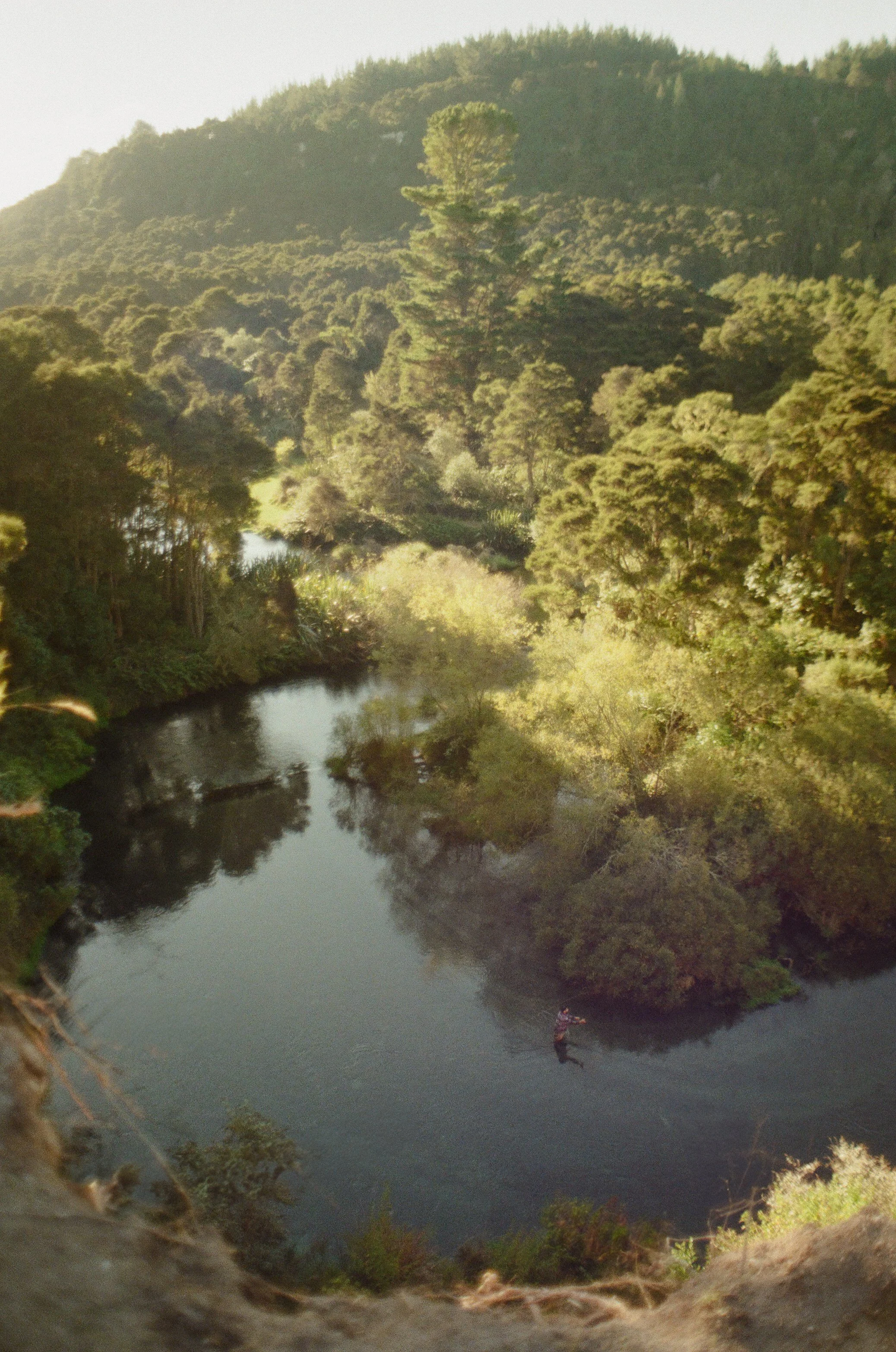 A person fishing in a calm river surrounded by dense green forest and hills.