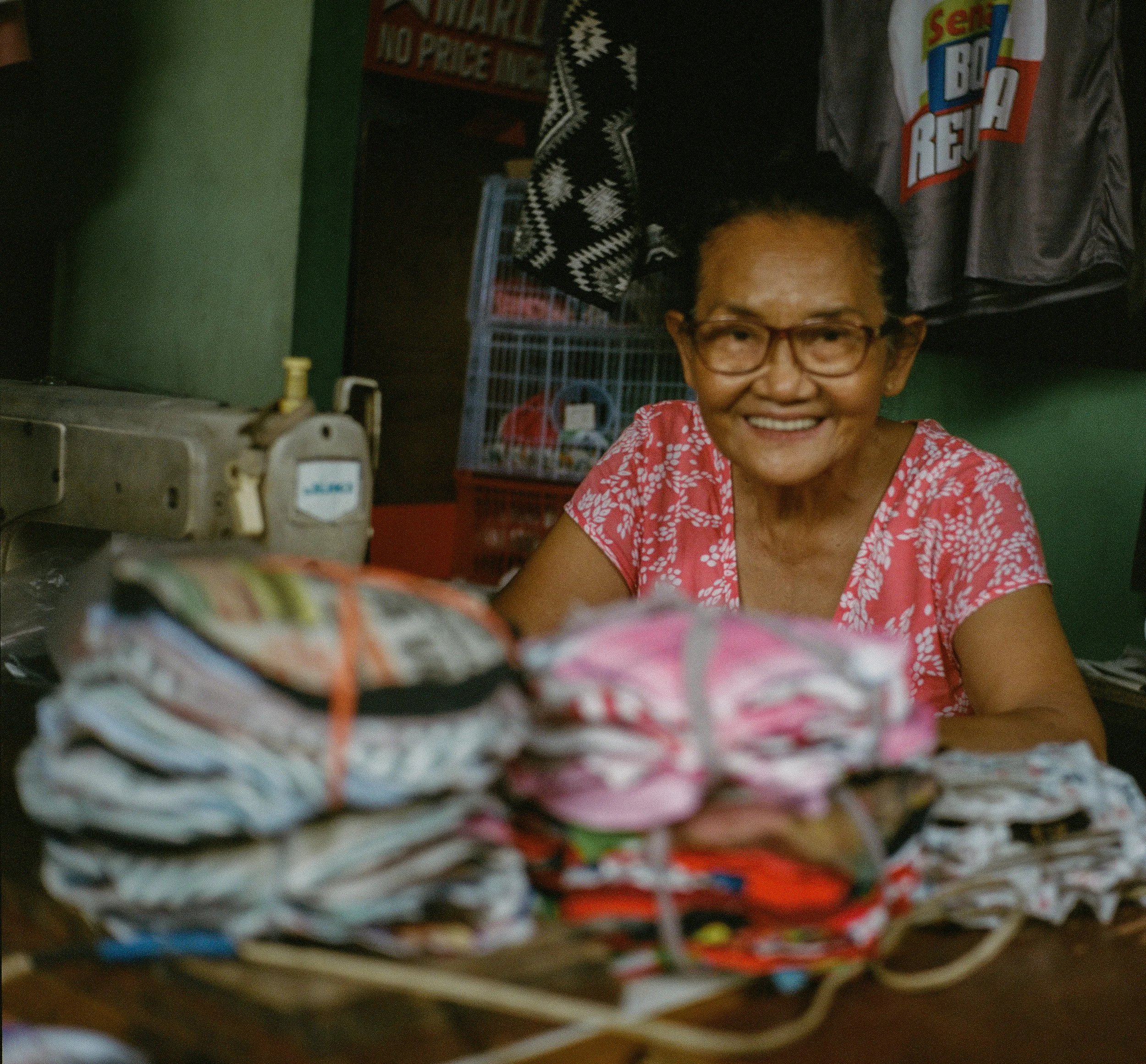 Smiling elderly woman with glasses behind a table of neatly folded colorful clothes in a small shop or stall.