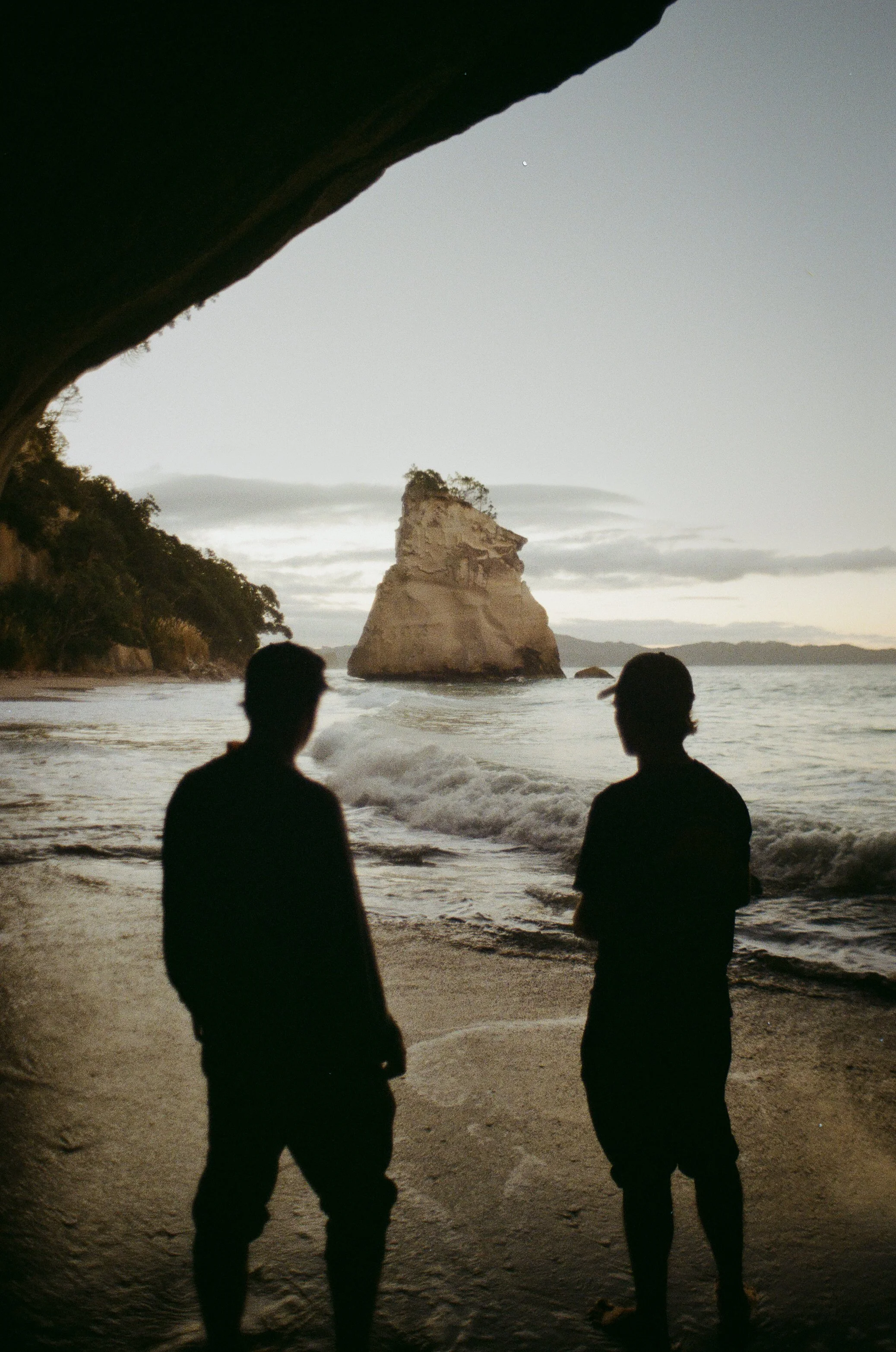 Silhouettes of two people standing on a beach, looking at the ocean and a large rock formation in the water during sunset or dusk, viewed from inside a cave or overhang.