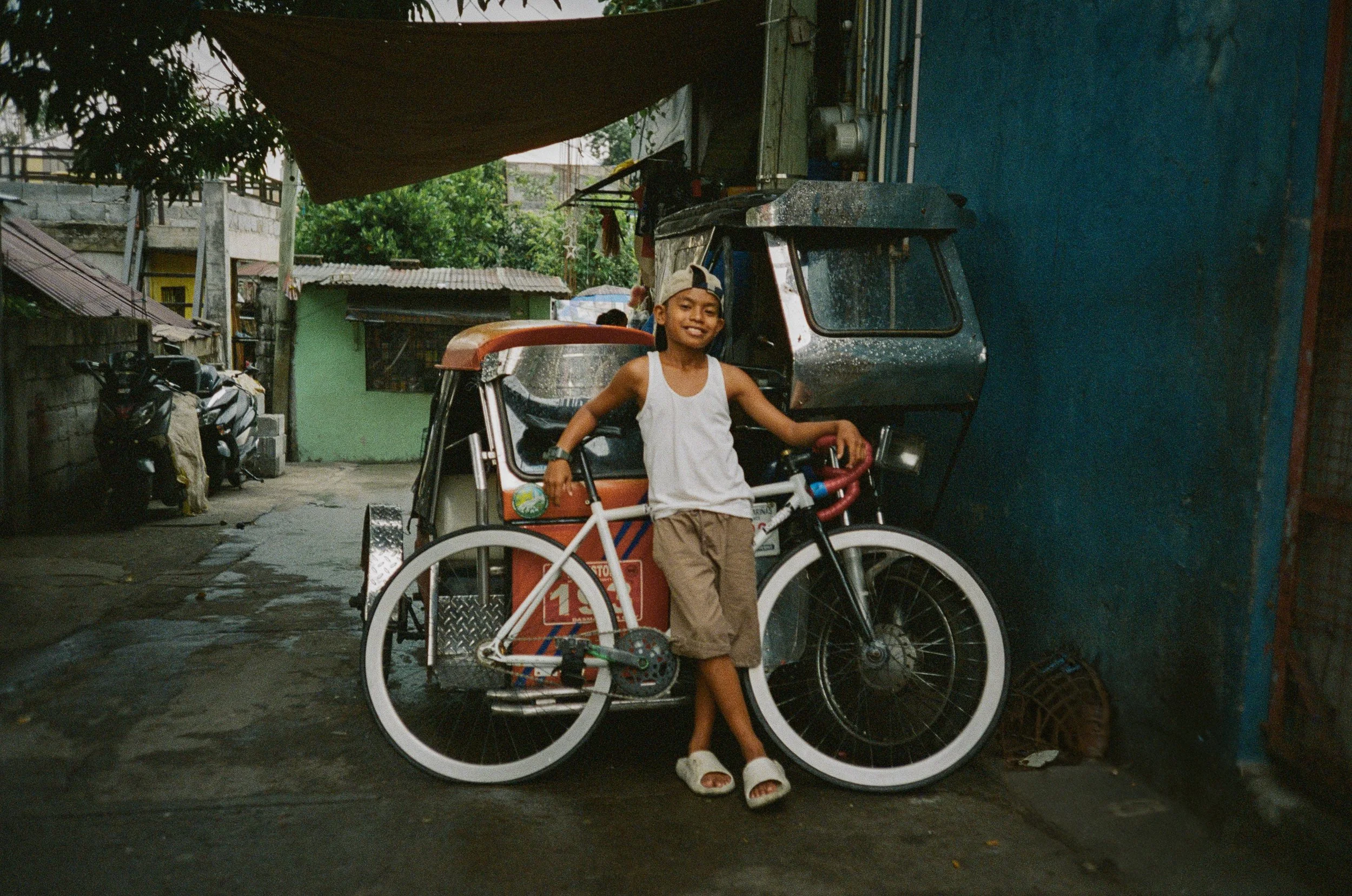 A young boy standing and smiling with one arm on a bicycle rickshaw in a narrow alley with motorcycles, small buildings, and greenery in the background.