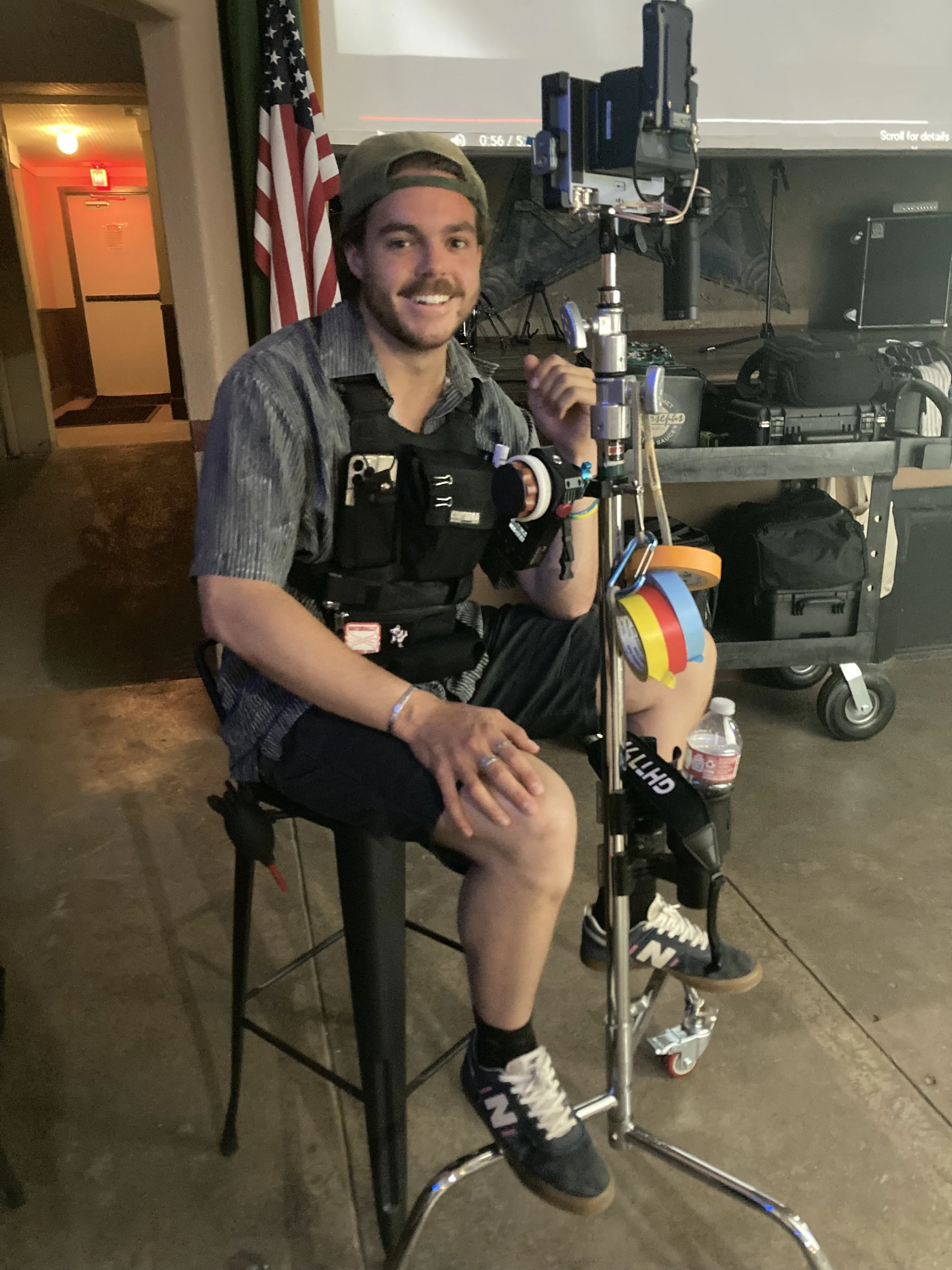 A man sitting on a stool next to camera equipment, wearing a vest with gear, sneakers, and a cap, smiling at the camera in a room with American flag and equipment on a cart.