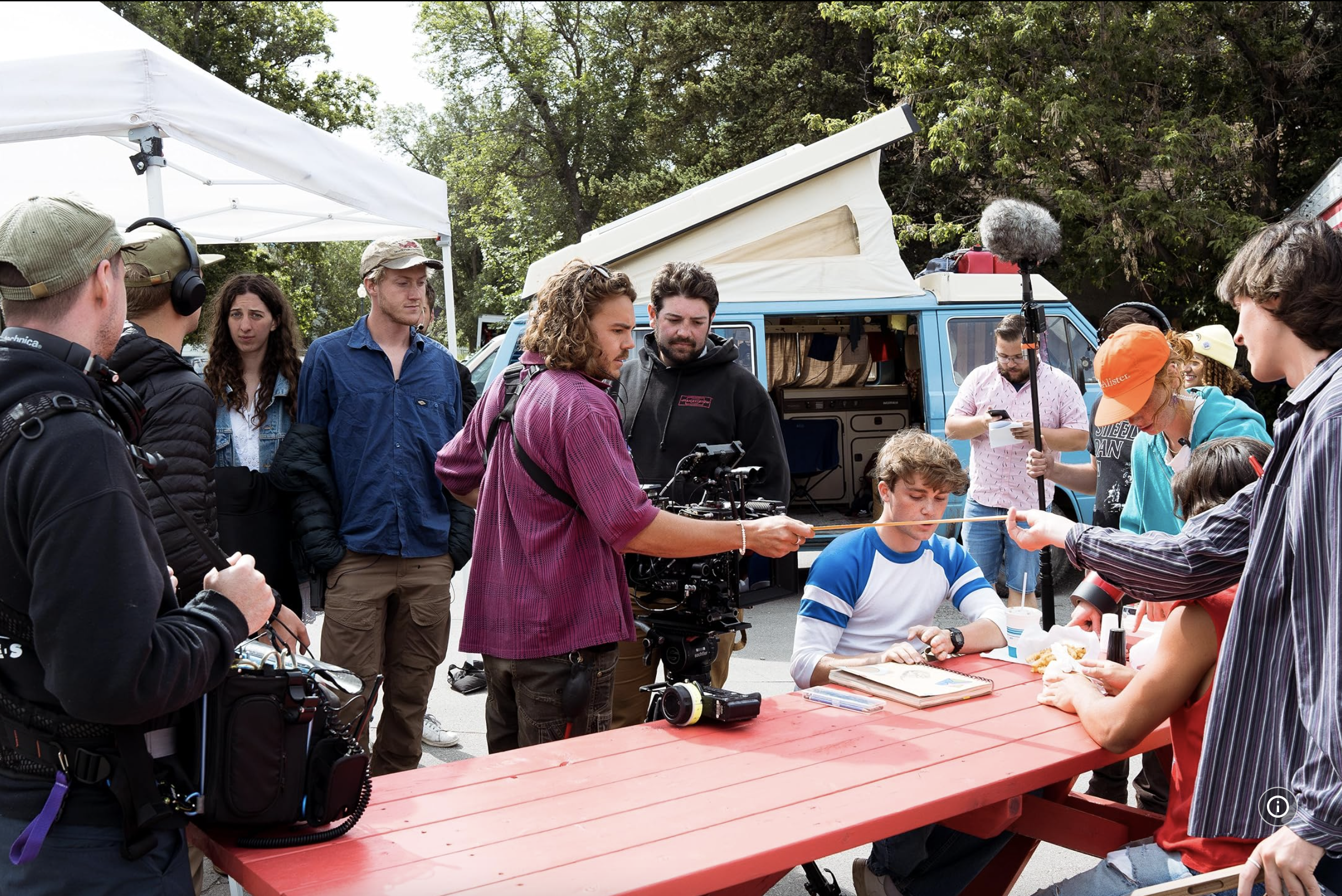 Filming crew and actors on a film set outdoors with a blue van, trees, and equipment.