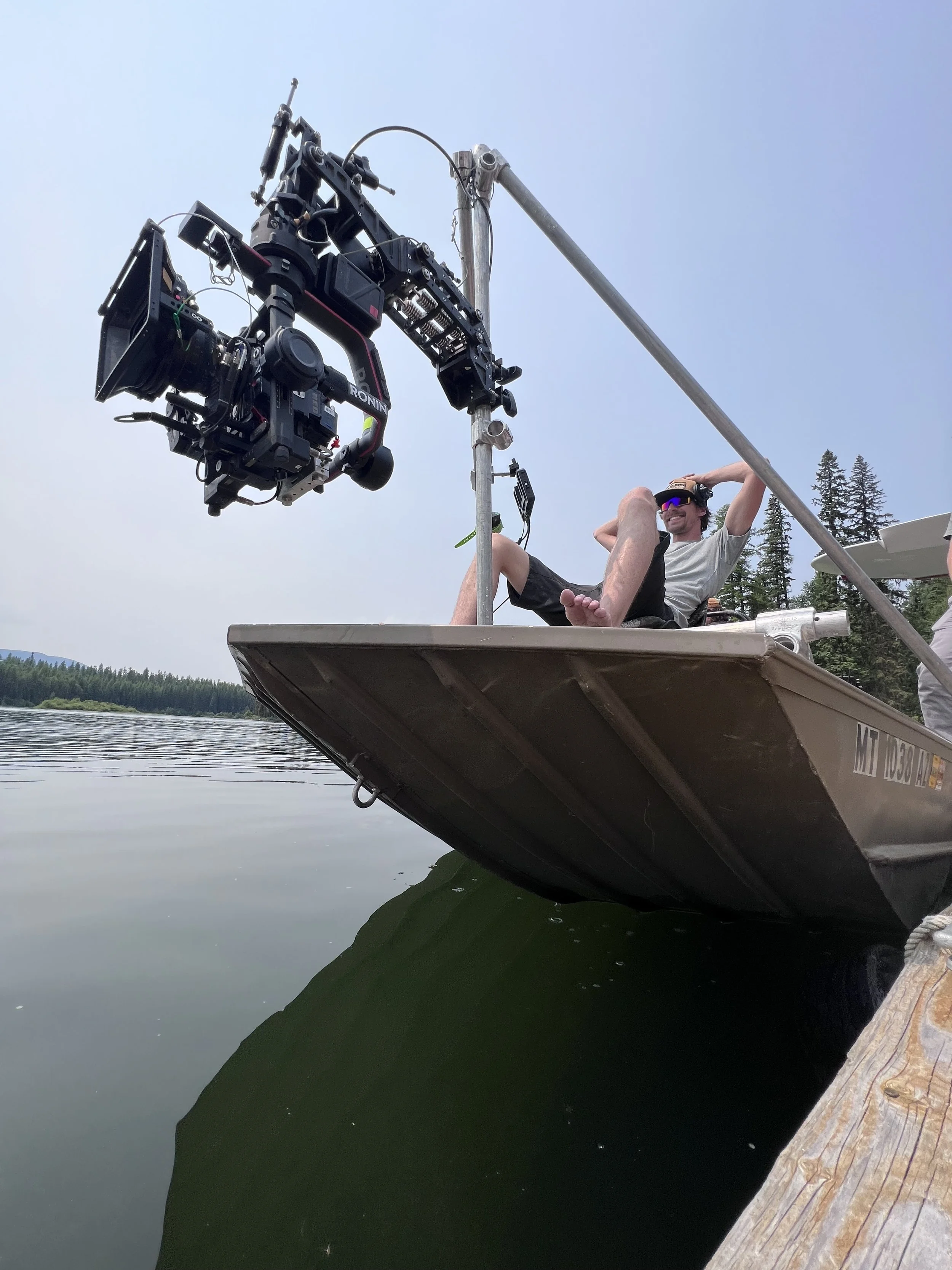 Person sitting on a boat with a camera crane, smiling, wearing sunglasses and headphones, on a calm lake with trees in the background