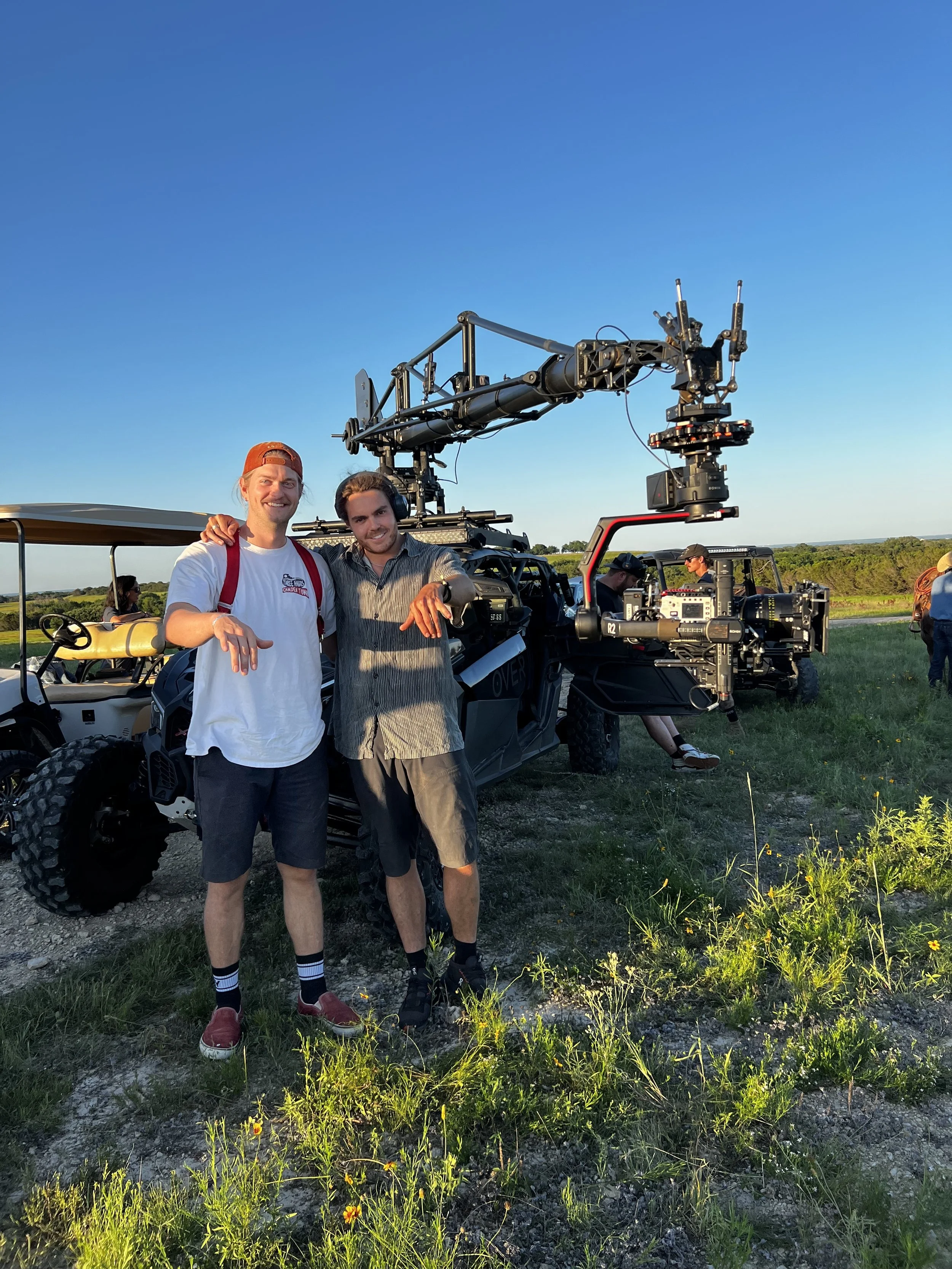 Two men standing in front of a large film camera on a tripod outdoors during sunset, smiling and pointing at the camera. The background features a grassy field and a clear blue sky.