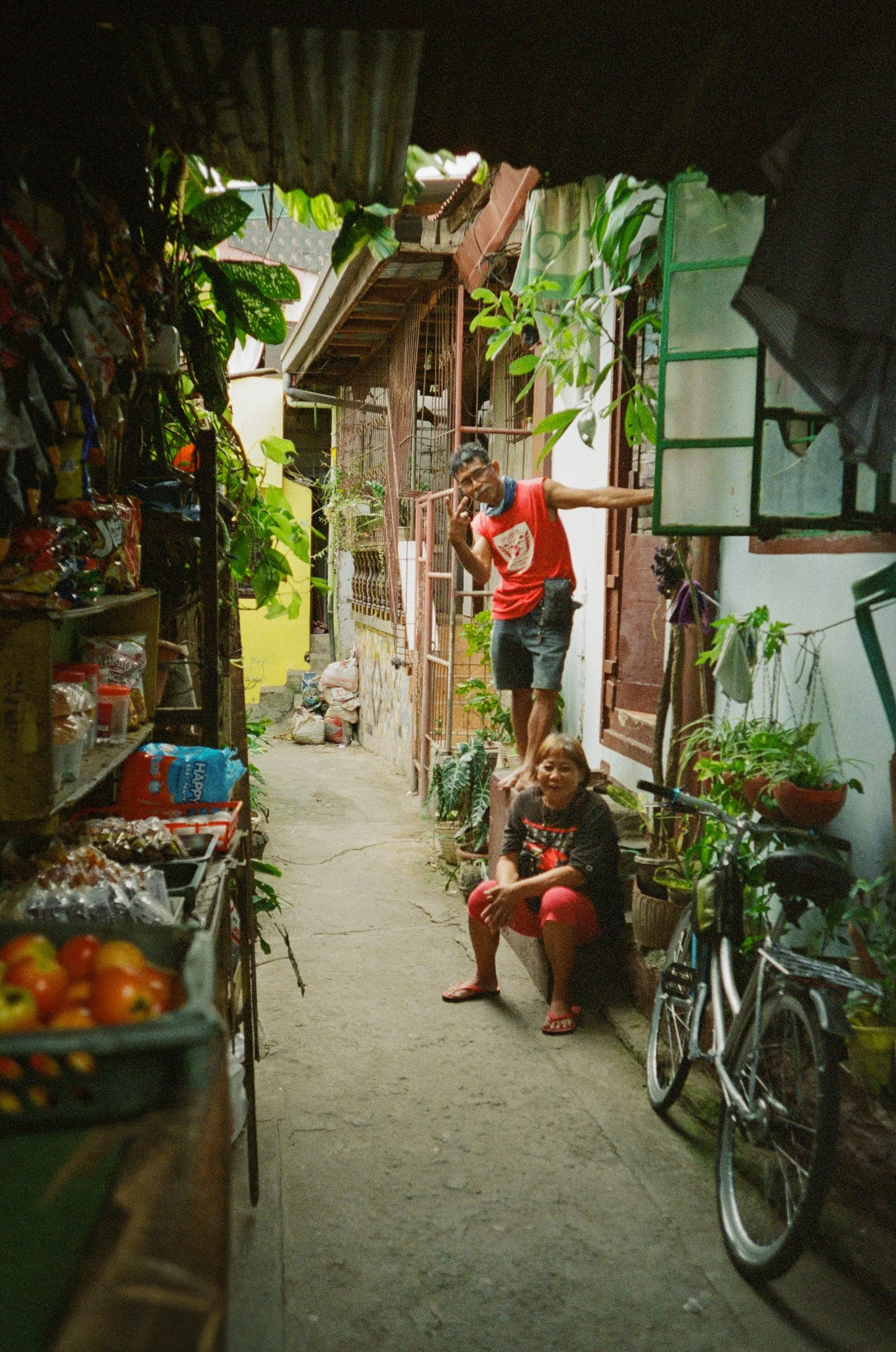 A man and a woman in an alleyway of a residential area, with plants, a bicycle, and various household items visible.