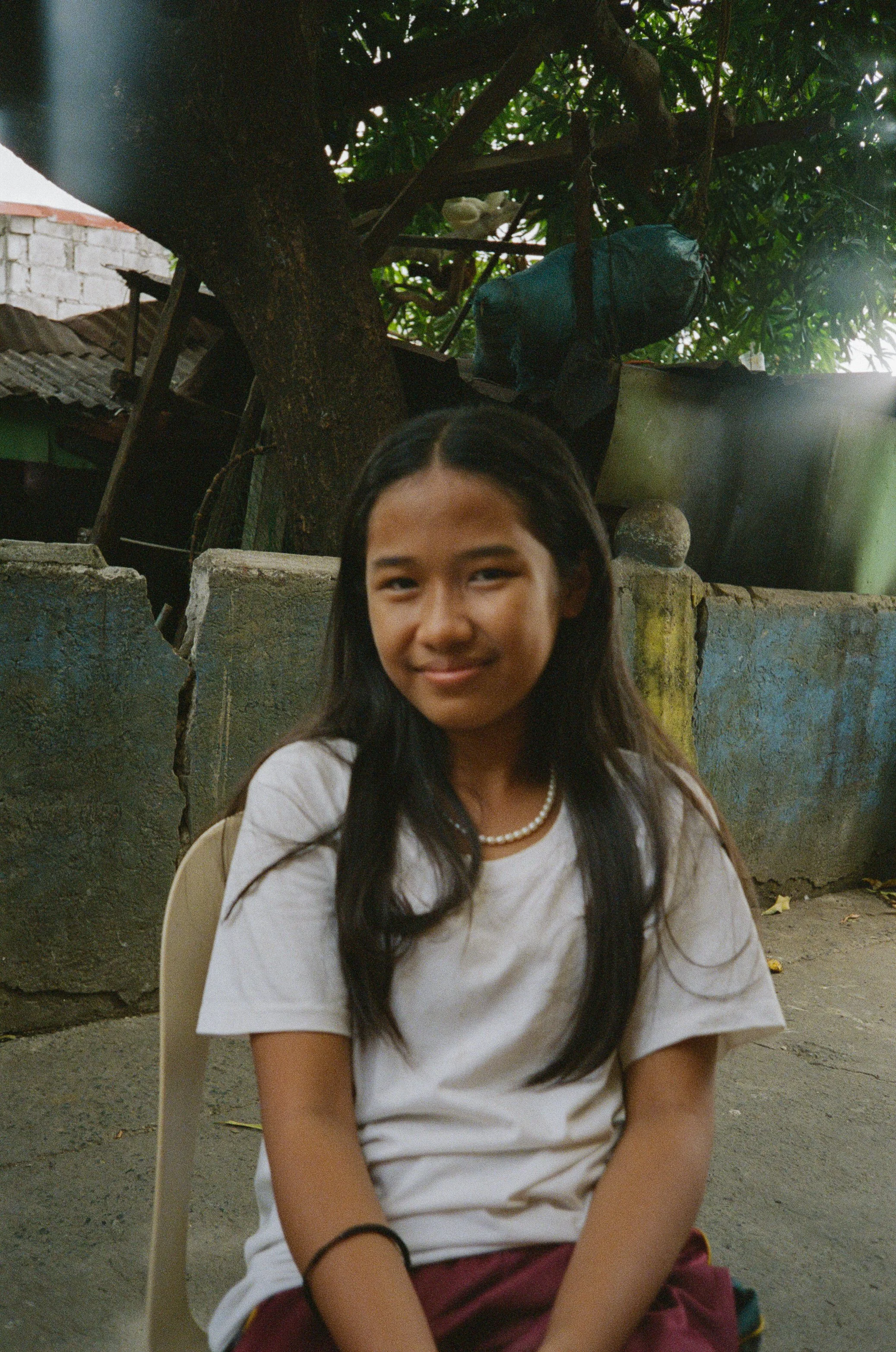 A young girl with long black hair, wearing a white t-shirt and a pearl necklace, sitting outdoors in front of a stone wall and tree, smiling at the camera.