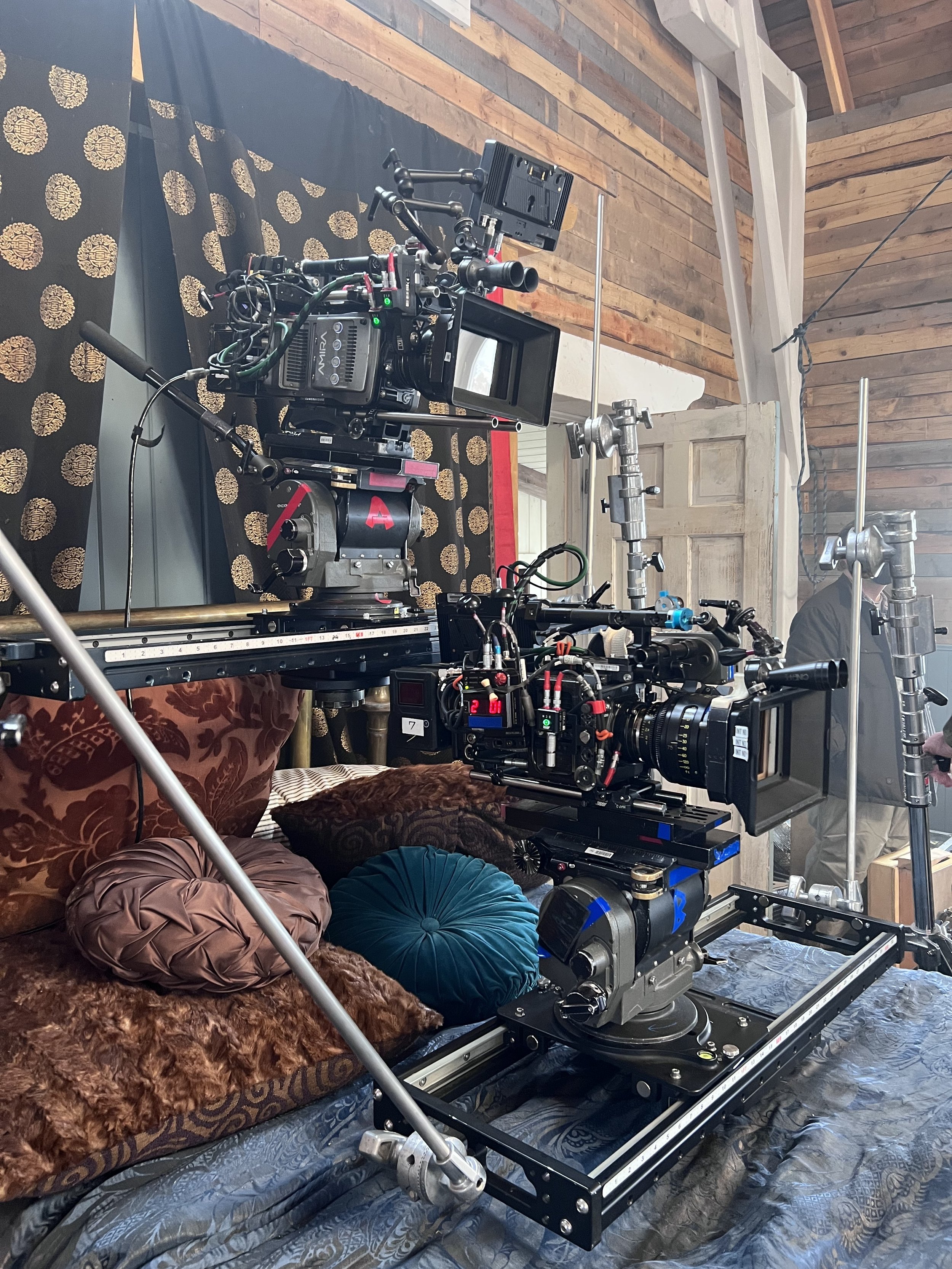 Close-up of professional camera equipment set up on a bed with decorative pillows, in a rustic room with wood-paneled walls and a patterned black backdrop.