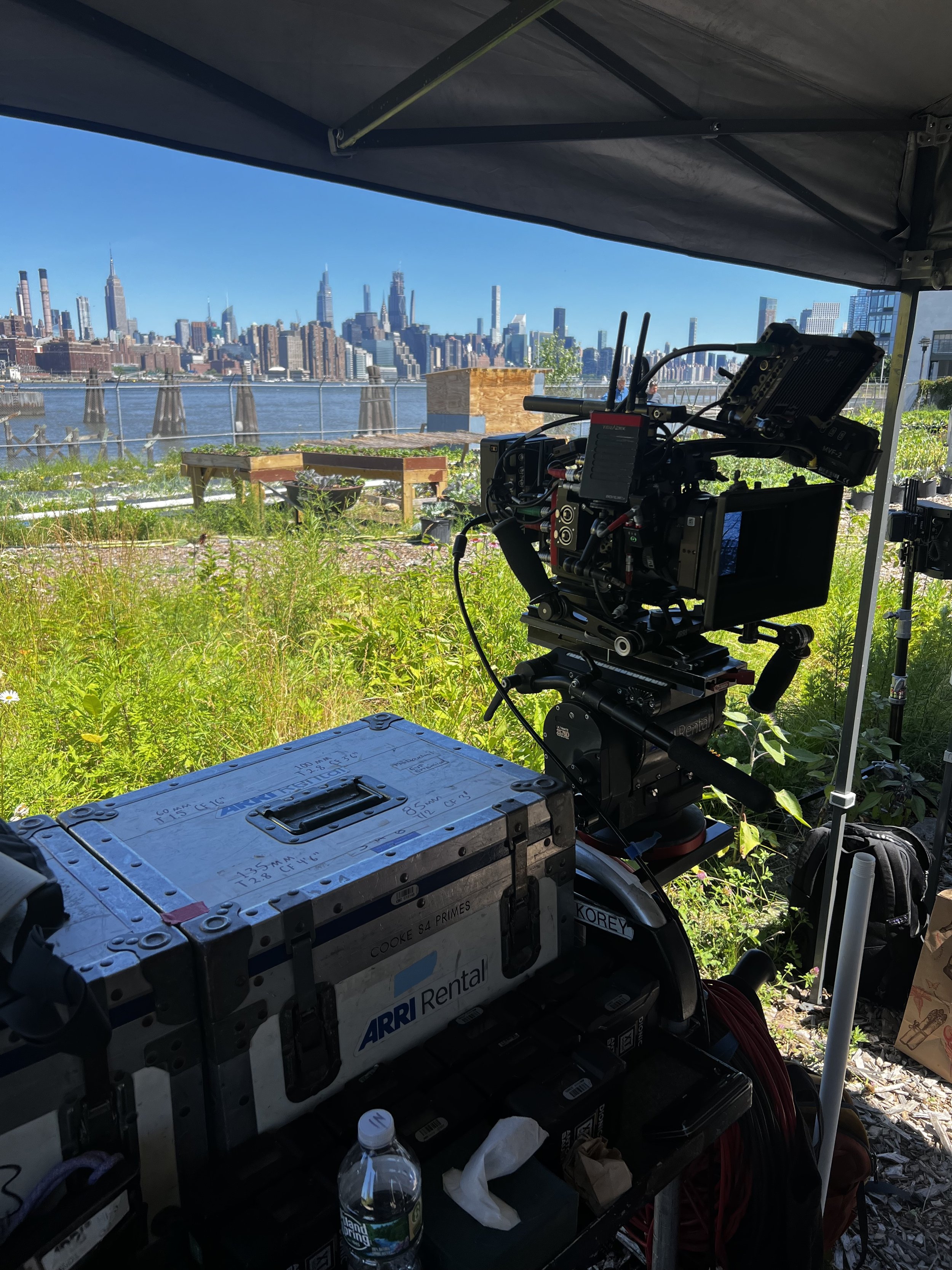 A film or photography set with a professional camera mounted on a tripod, positioned under a canopy, overlooking a city skyline across a river, with green vegetation and some equipment and supplies in the foreground.