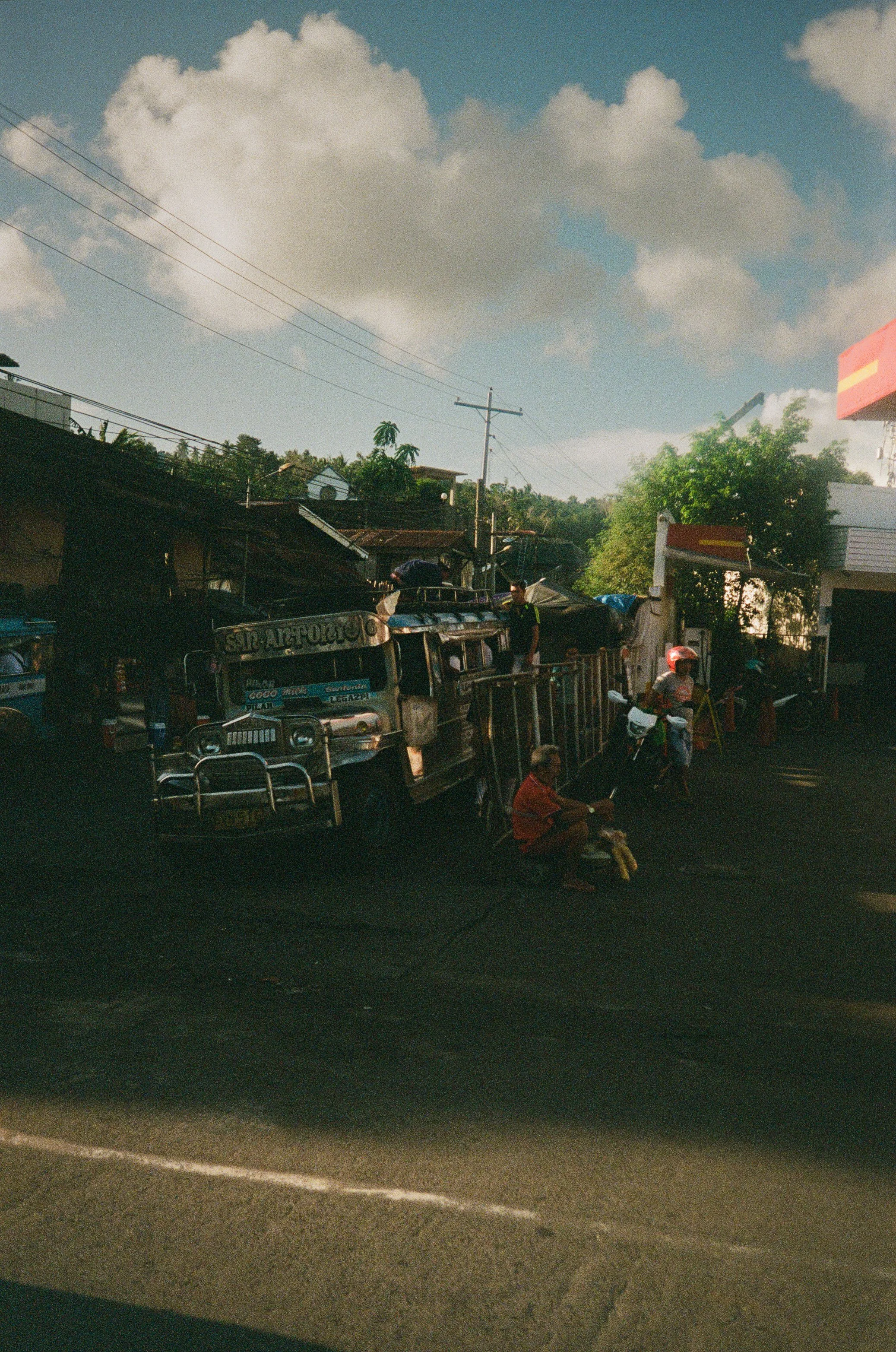 Street scene showing a jeepney, a motorcycle, a person sitting on the curb reading, and several pedestrians on a sunny day with scattered clouds.