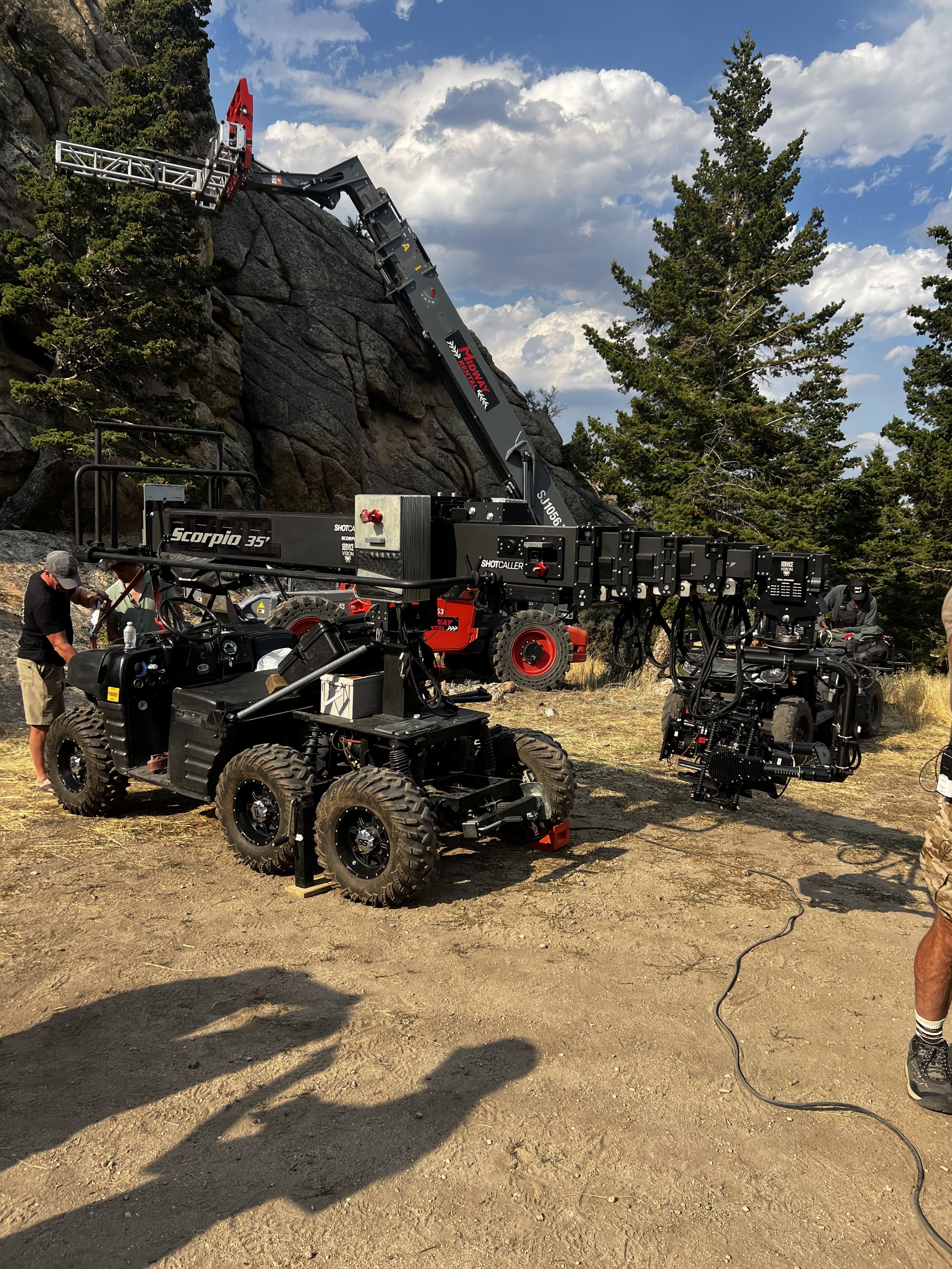 A camera crane rig set up outdoors near a rocky hillside and trees, with crew members working around it on a sunny day.