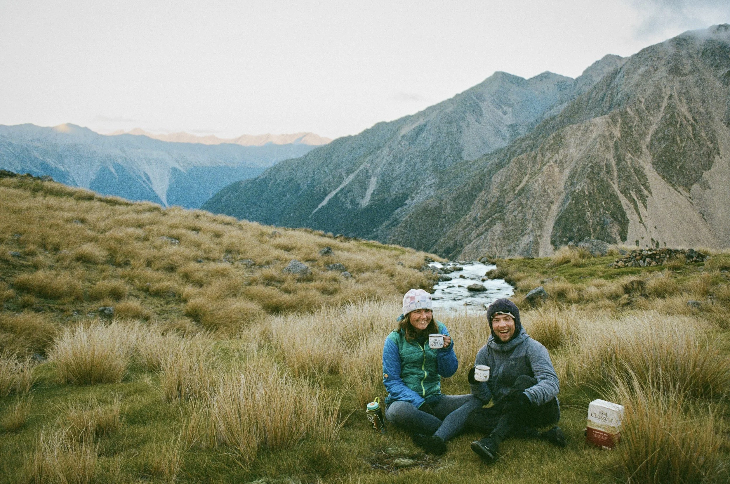 Two people sitting on grass in a mountainous landscape, smiling and holding cups, with a river running through the valley behind them.