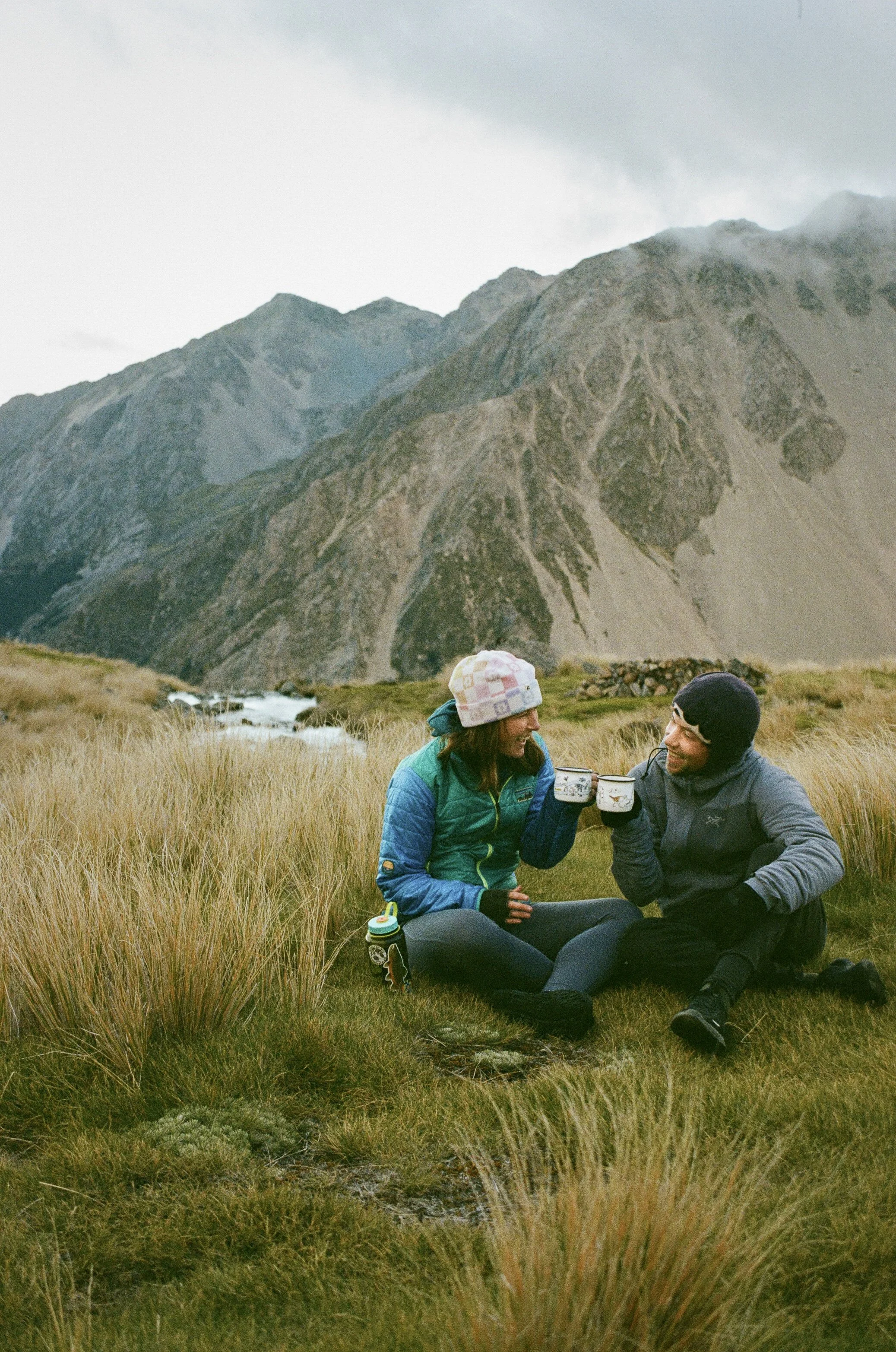 Two people sitting on grass in a mountainous area, holding mugs and smiling at each other, with cloudy sky and rugged mountains in the background.