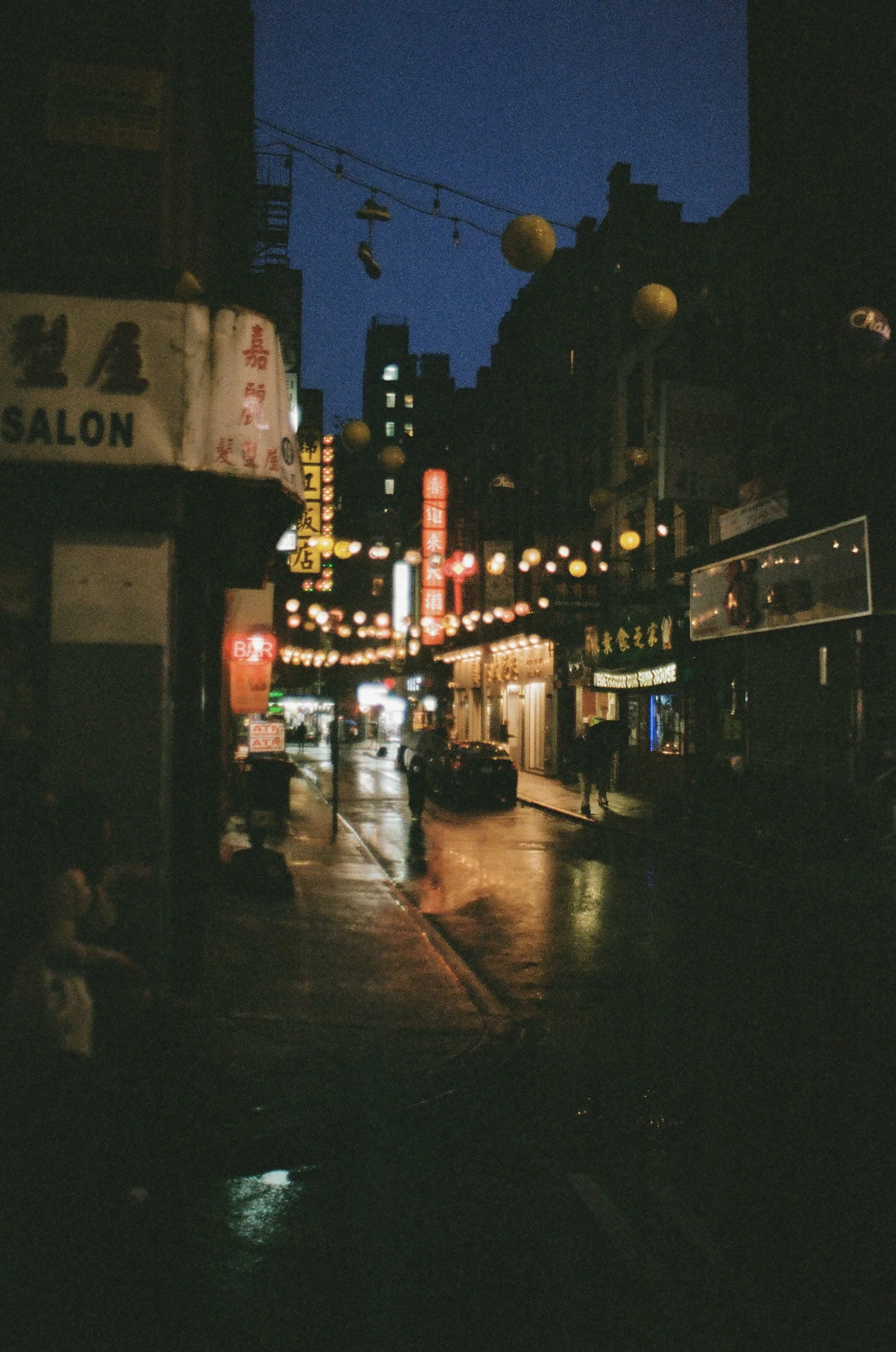 A dimly lit city street at night, with hanging lanterns, neon signs, and a few pedestrians.