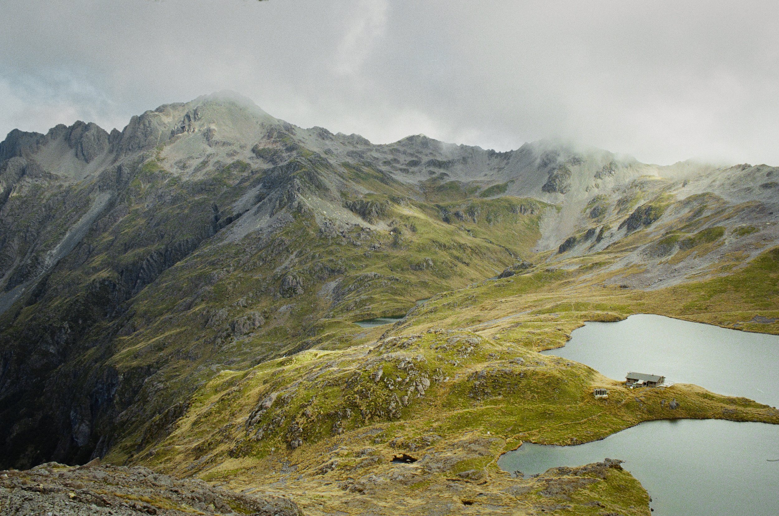 Mountain landscape with steep, rocky peaks, grassy slopes, and two lakes in the foreground, partially shrouded in mist and clouds.