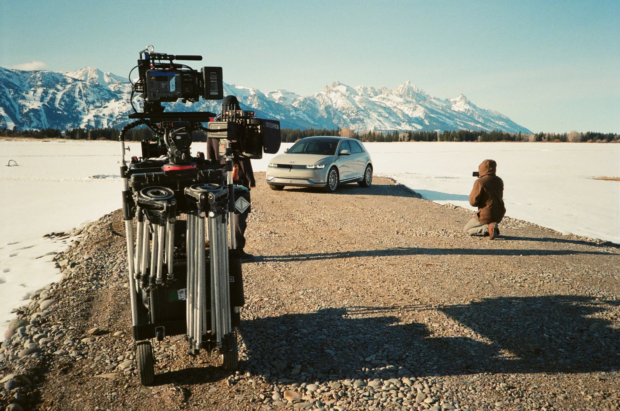 A behind-the-scenes look at a film or photo shoot in a snowy outdoor landscape with mountains in the background. The setup includes a camera on a tripod, a photographer kneeling on the ground taking pictures of a silver car, and a crew member standin
