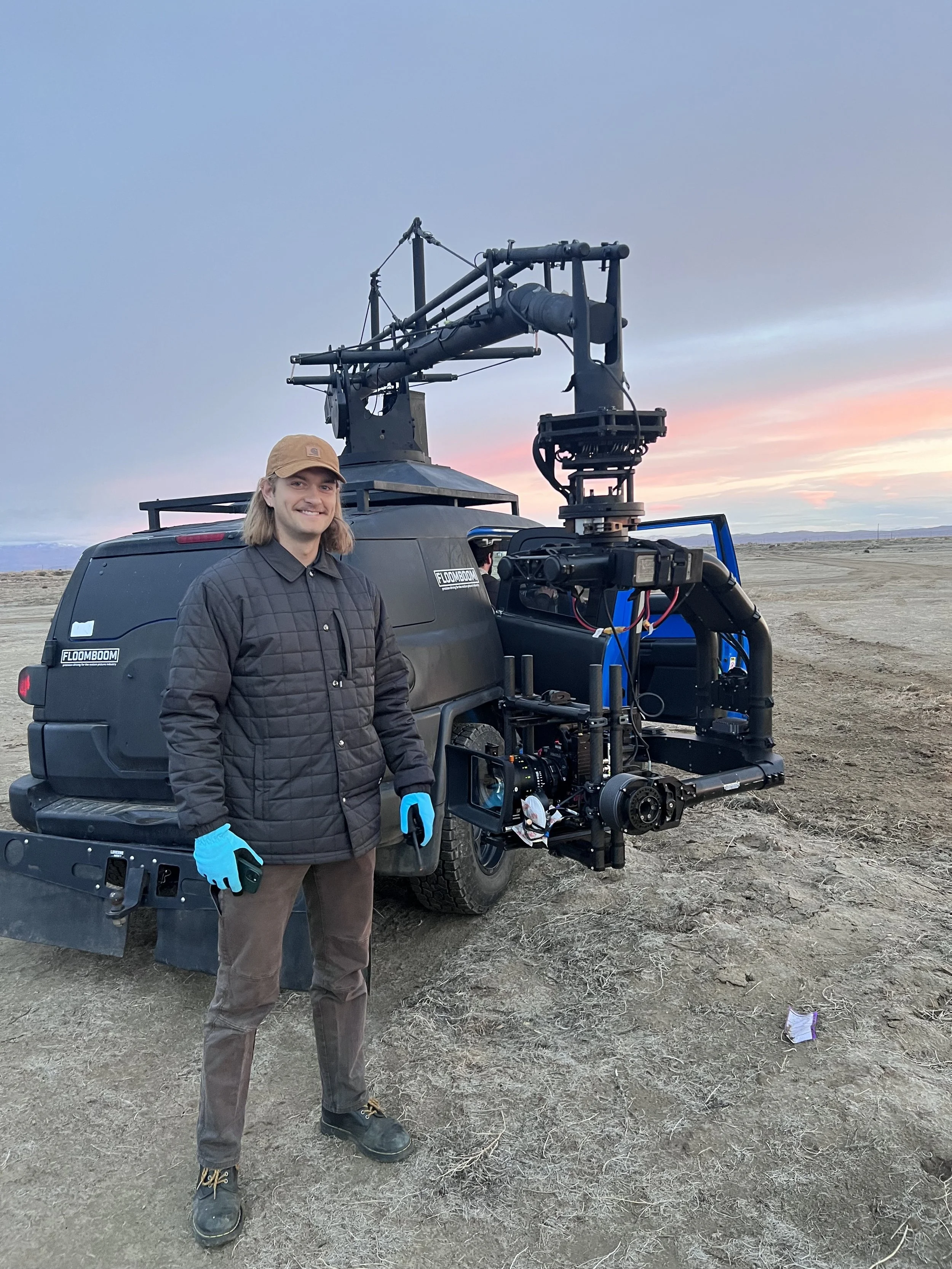 A man wearing a brown cap, black quilted jacket, blue gloves, and brown pants standing in front of a large vehicle with a camera rig attached. The background shows a flat outdoor landscape at sunset.