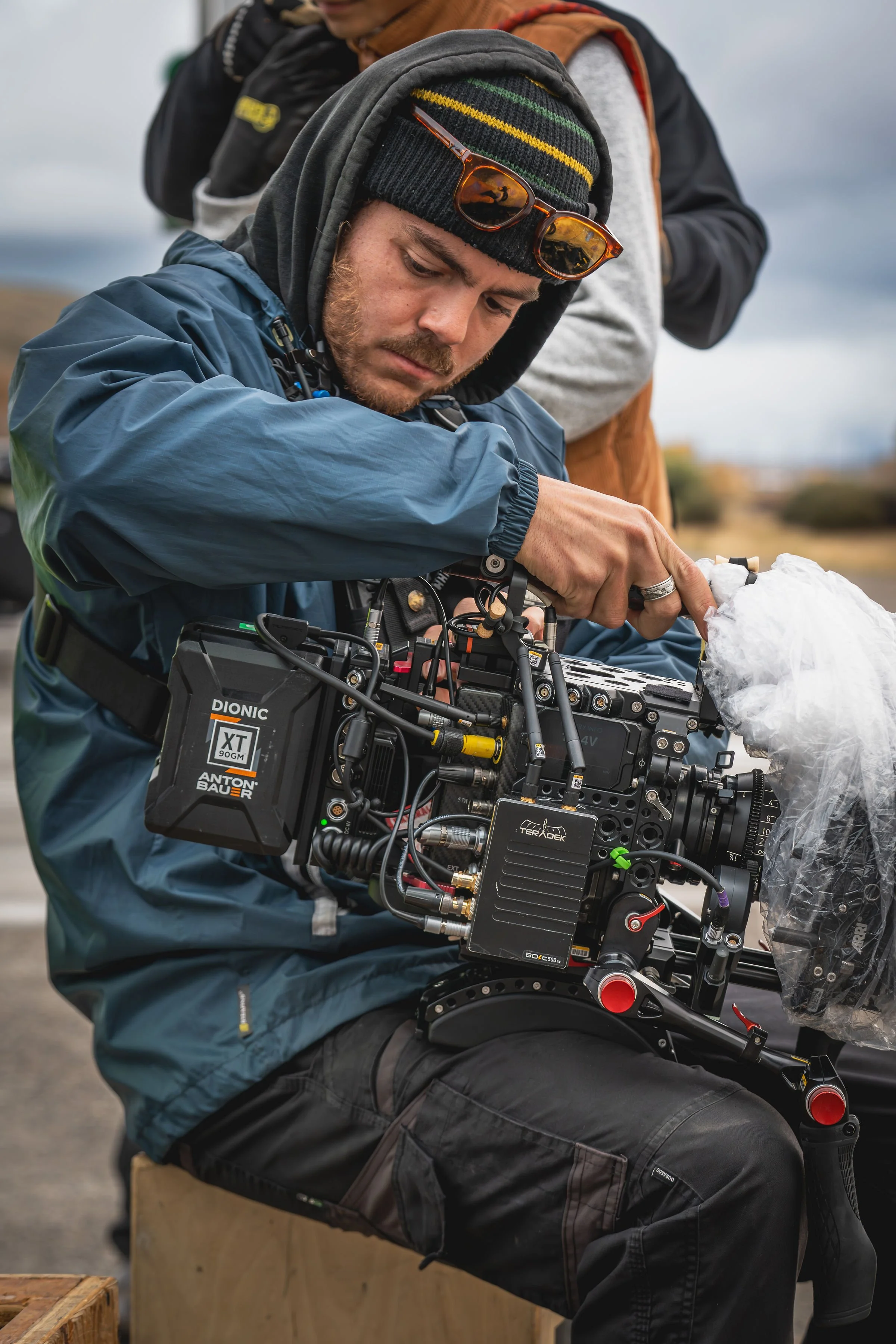 A man operating a professional film camera outdoors, wearing a blue jacket, a gray hoodie, a beanie with colorful stripes, and sunglasses resting on his head.