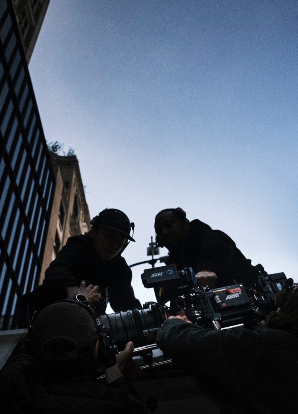 Filmmakers work with camera equipment outdoors during dusk, silhouetted against a twilight sky, with urban buildings in the background.