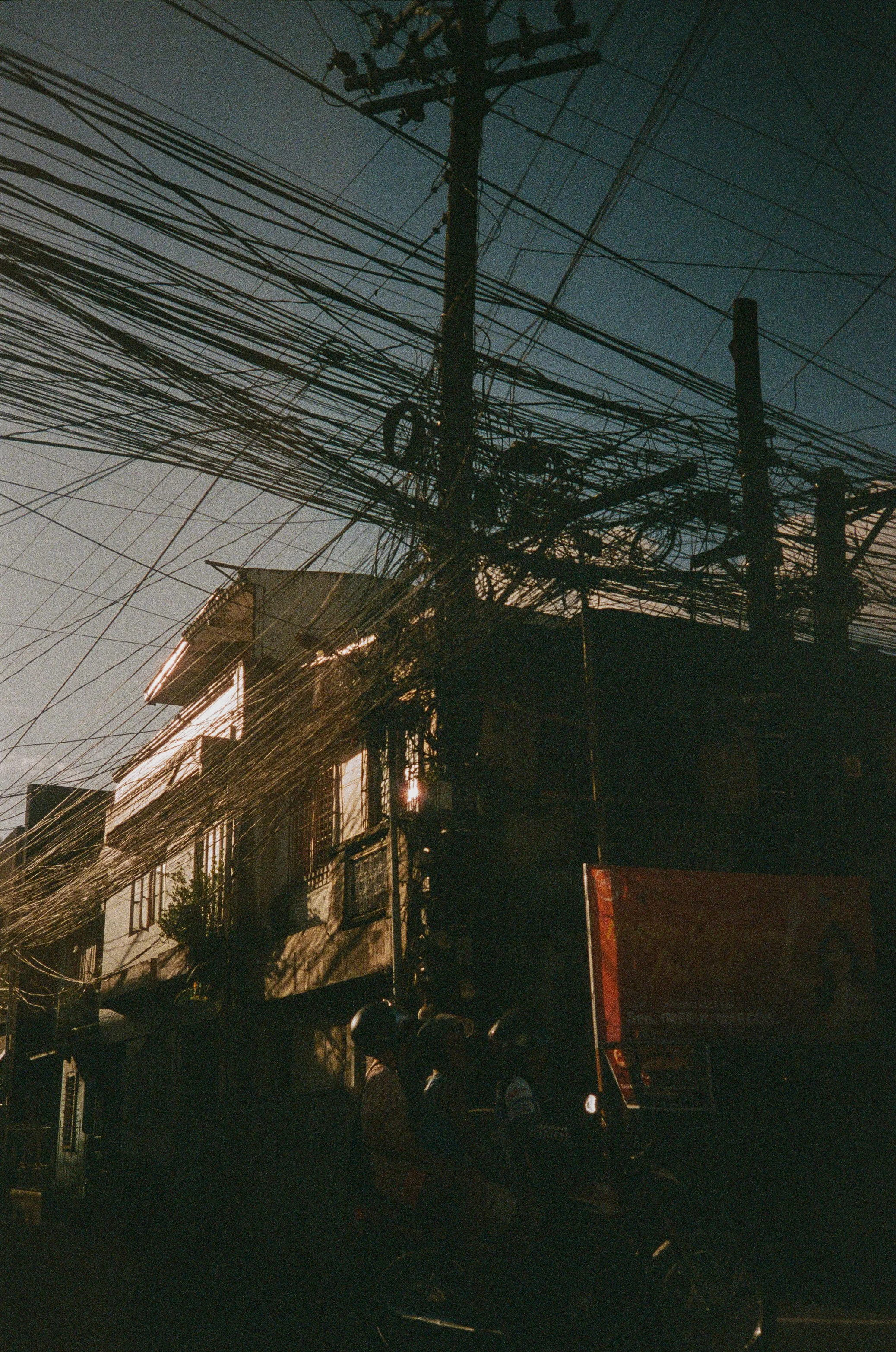 Street view with tangled power lines and electric poles at sunset, with silhouettes of people and buildings.
