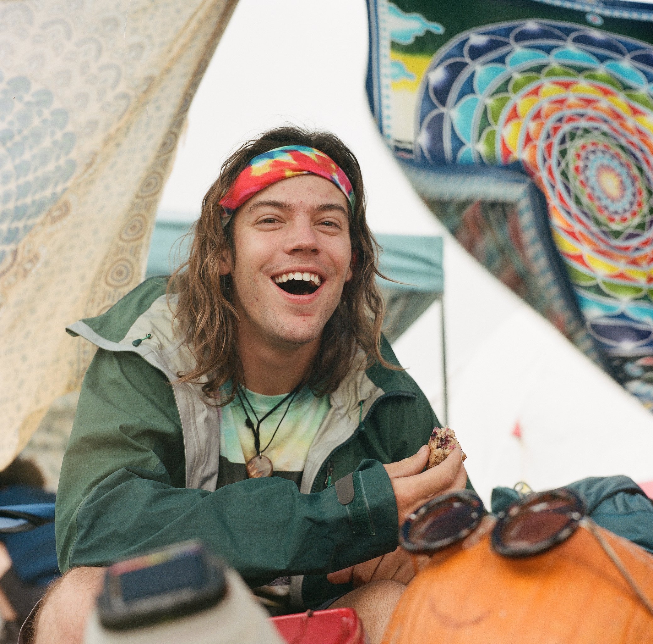 A young man with long hair, wearing a colorful headband, a tie-dye shirt, and a green jacket, smiling and holding a cookie at an outdoor gathering surrounded by colorful patterned blankets or tapestries and camping gear.