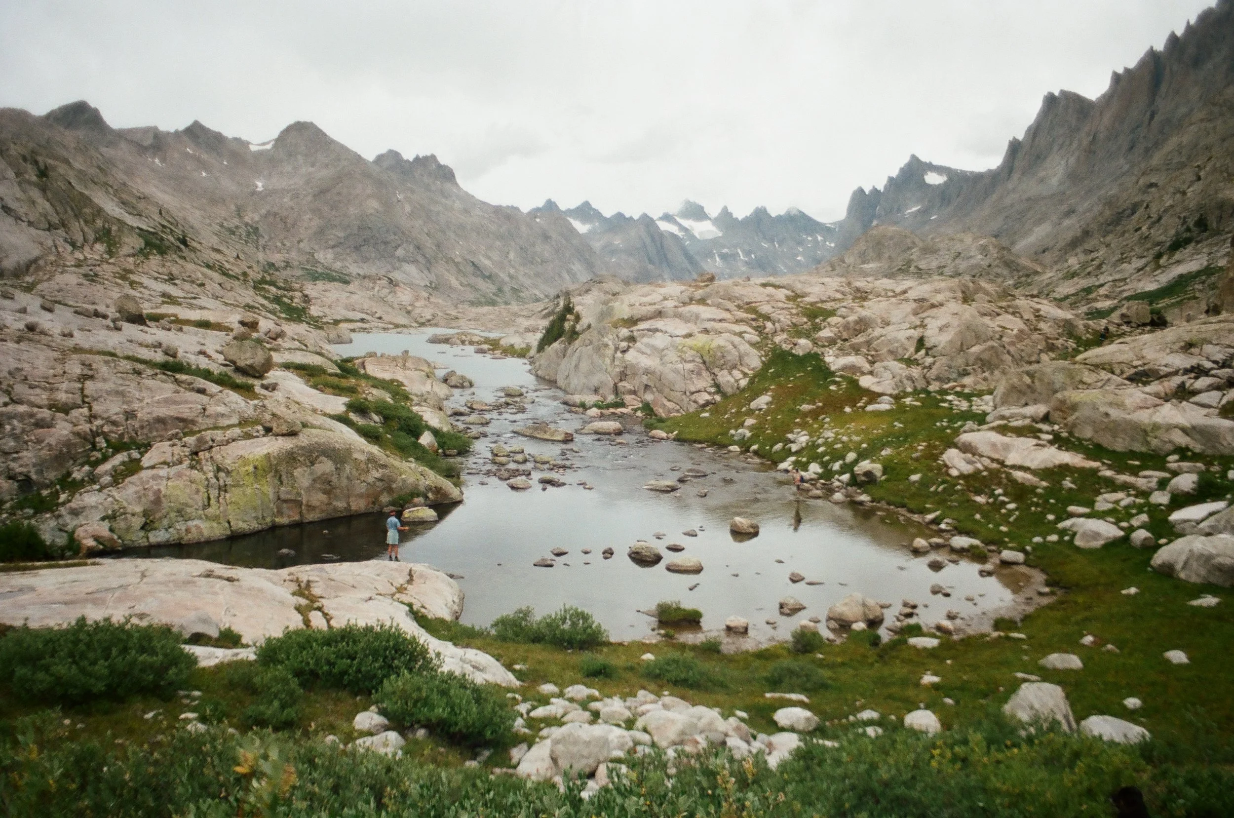 A mountainous landscape with a river running through the valley, surrounded by large rocks and sparse green vegetation, under an overcast sky.