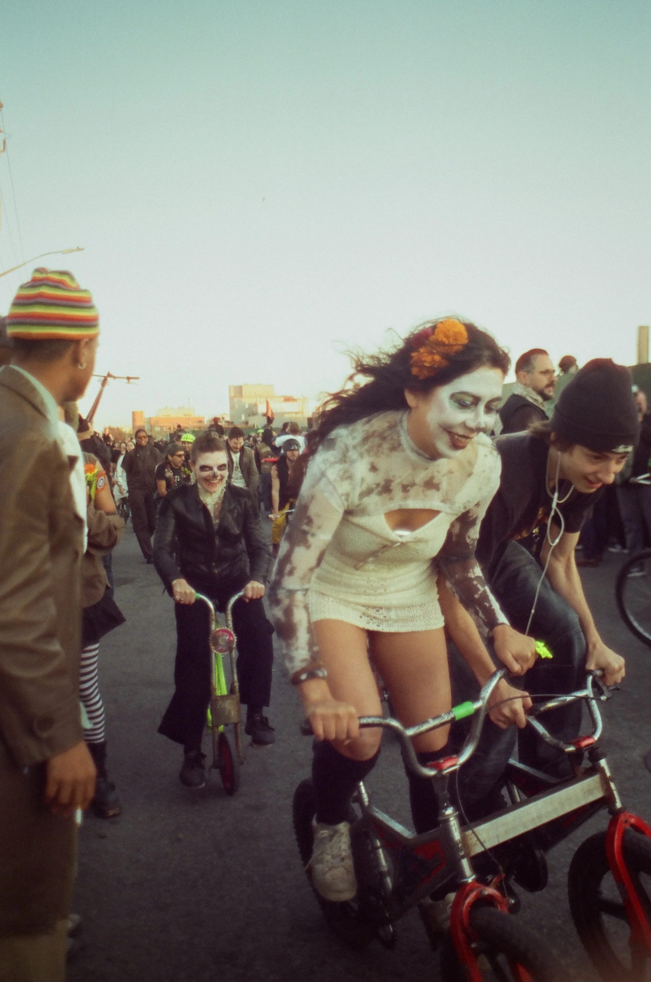 People dressed as zombies riding bikes and walking in a parade or event, with a crowd in the background and buildings in the distance.