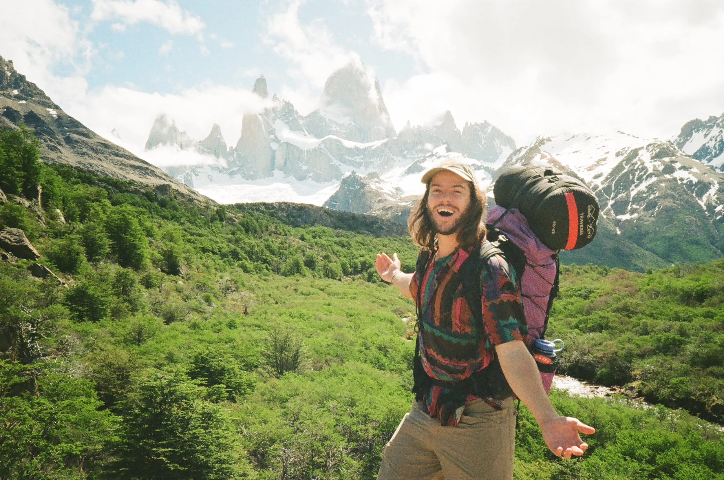 A smiling man with long hair and a beard, wearing a colorful shirt, tan shorts, and a cap, standing in a lush green valley with snow-capped mountains in the background, carrying a large backpack.
