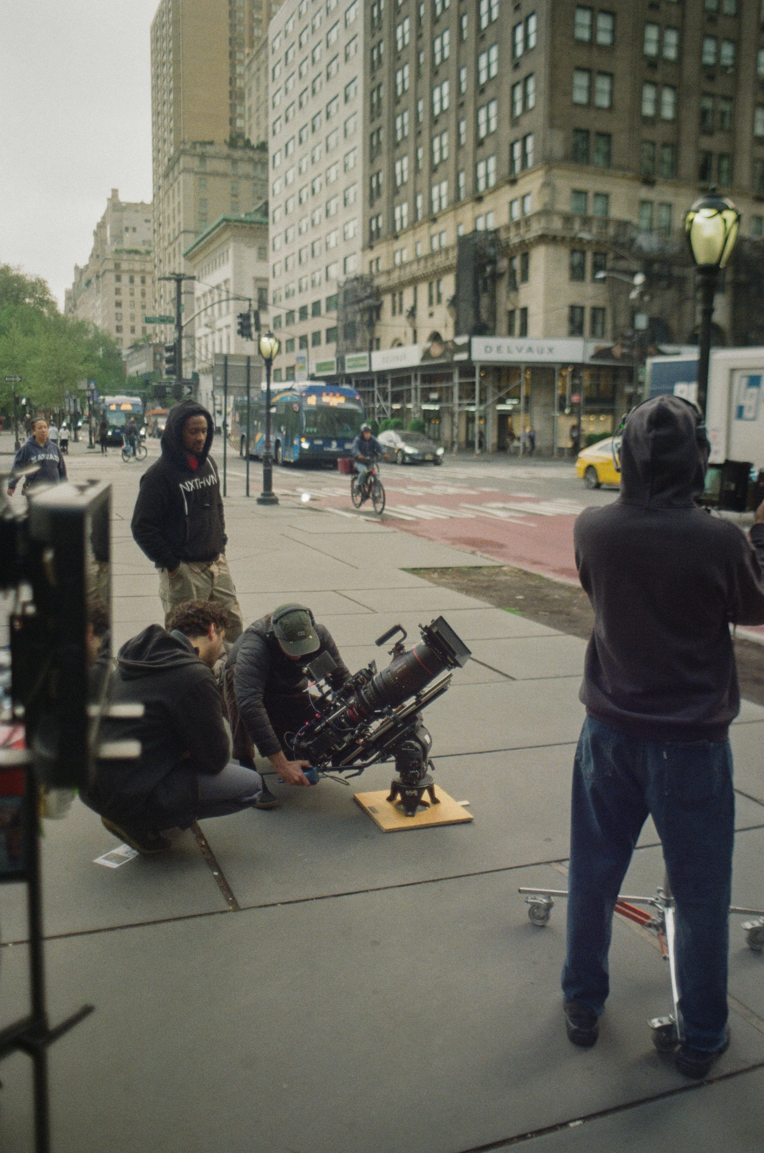 Filming crew setting up a camera on a city sidewalk with people and tall buildings in the background.