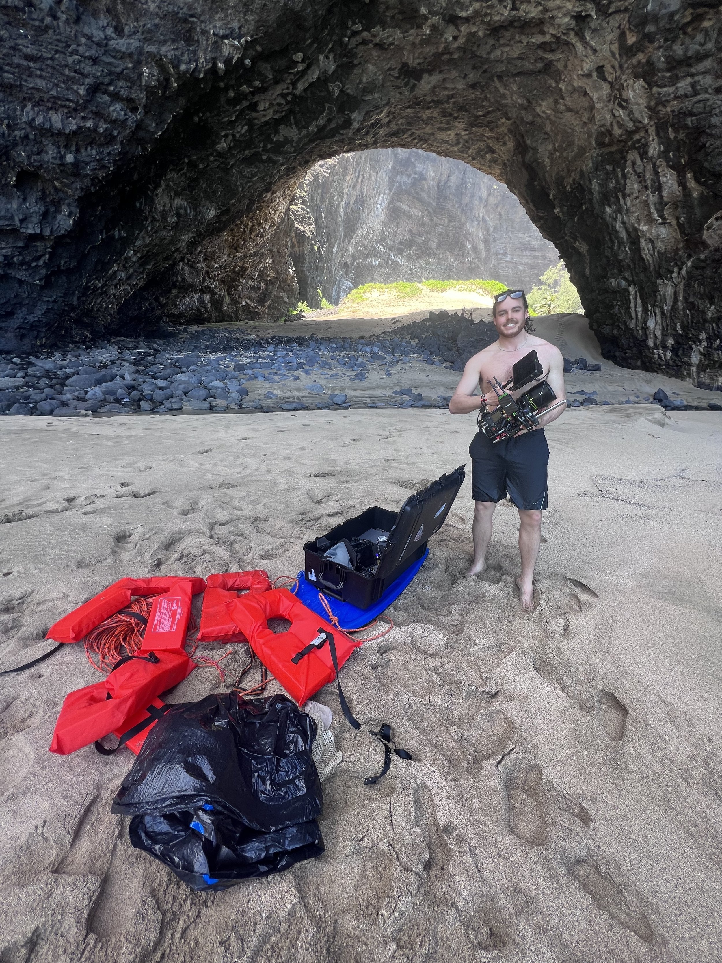 A man standing on the beach inside a rocky cave holding a camera rig, with equipment and life jackets laid out on the sand.