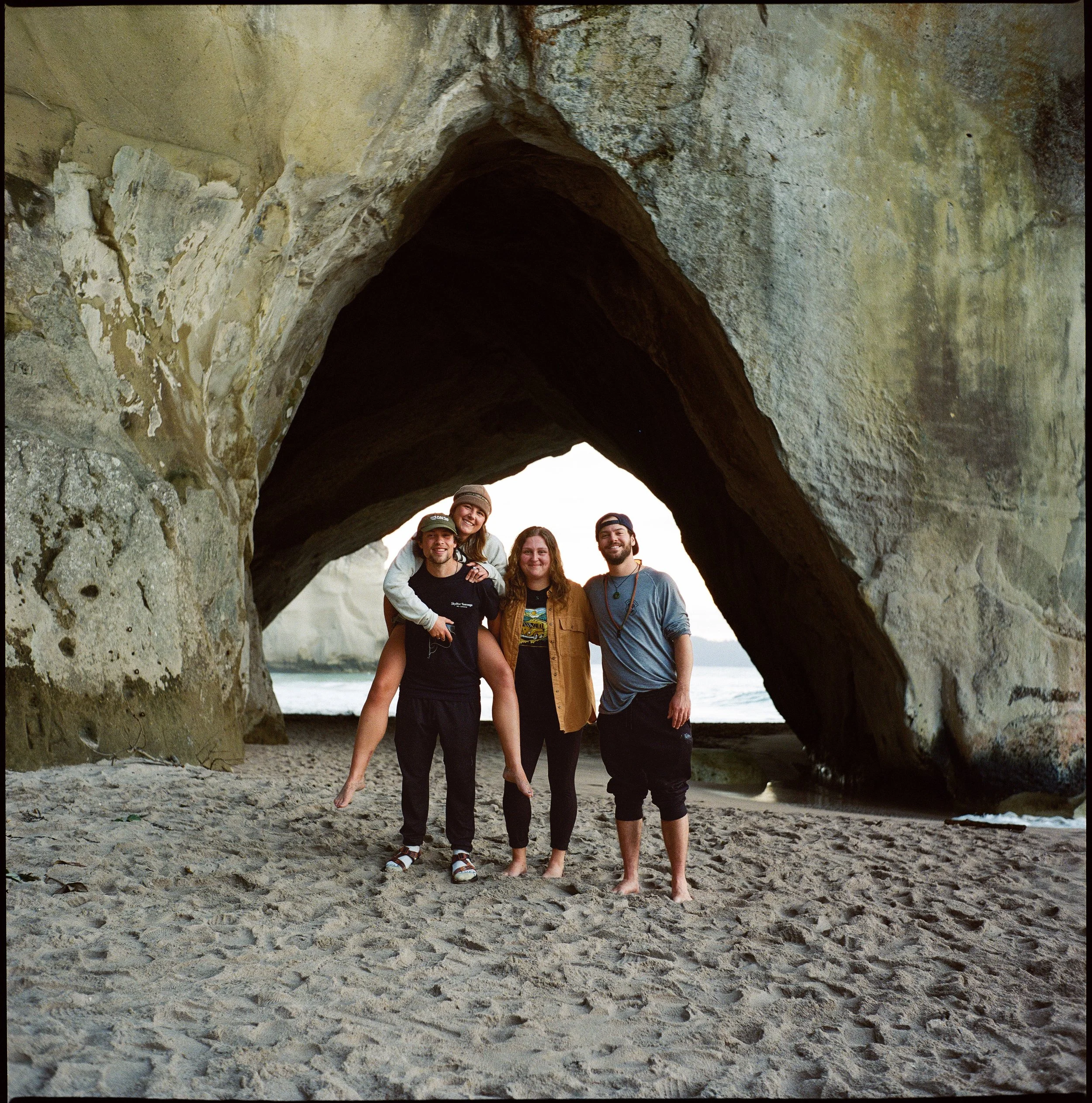 Four friends standing on the sandy beach beneath a large natural arch rock formation at the coast, smiling at the camera.