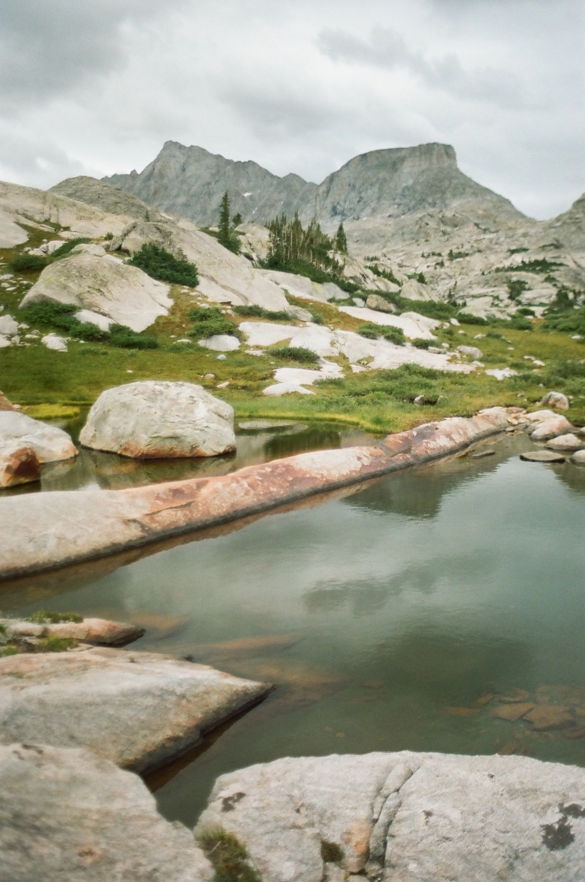 Mountain landscape with rocky terrain, green vegetation, and a pond in the foreground under a cloudy sky.