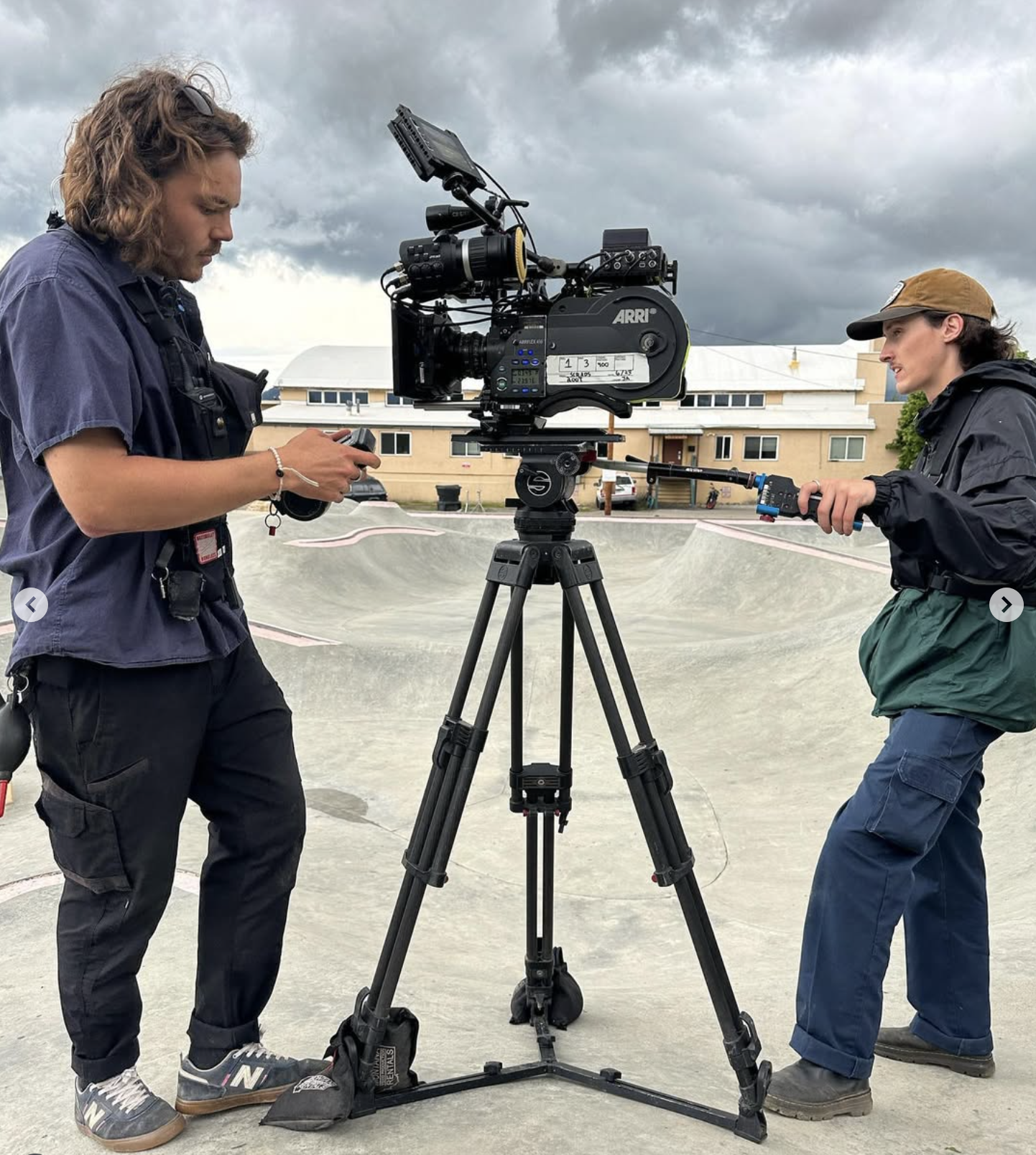 Two film crew members setting up a professional camera on a tripod outdoors, with a gray cloudy sky and a residential building in the background.