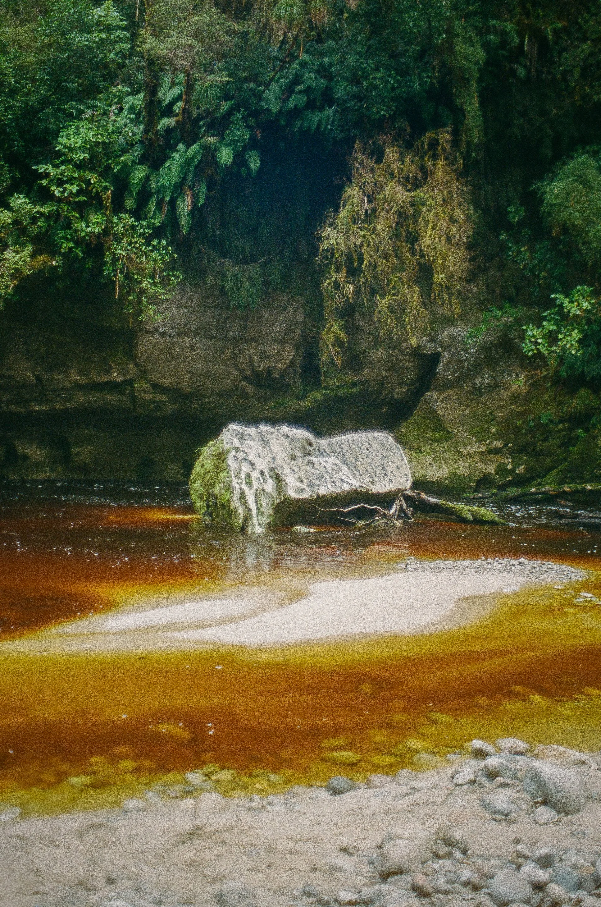 A river flowing through a lush, green forest with large moss-covered rocks and a sandy beach in the foreground.