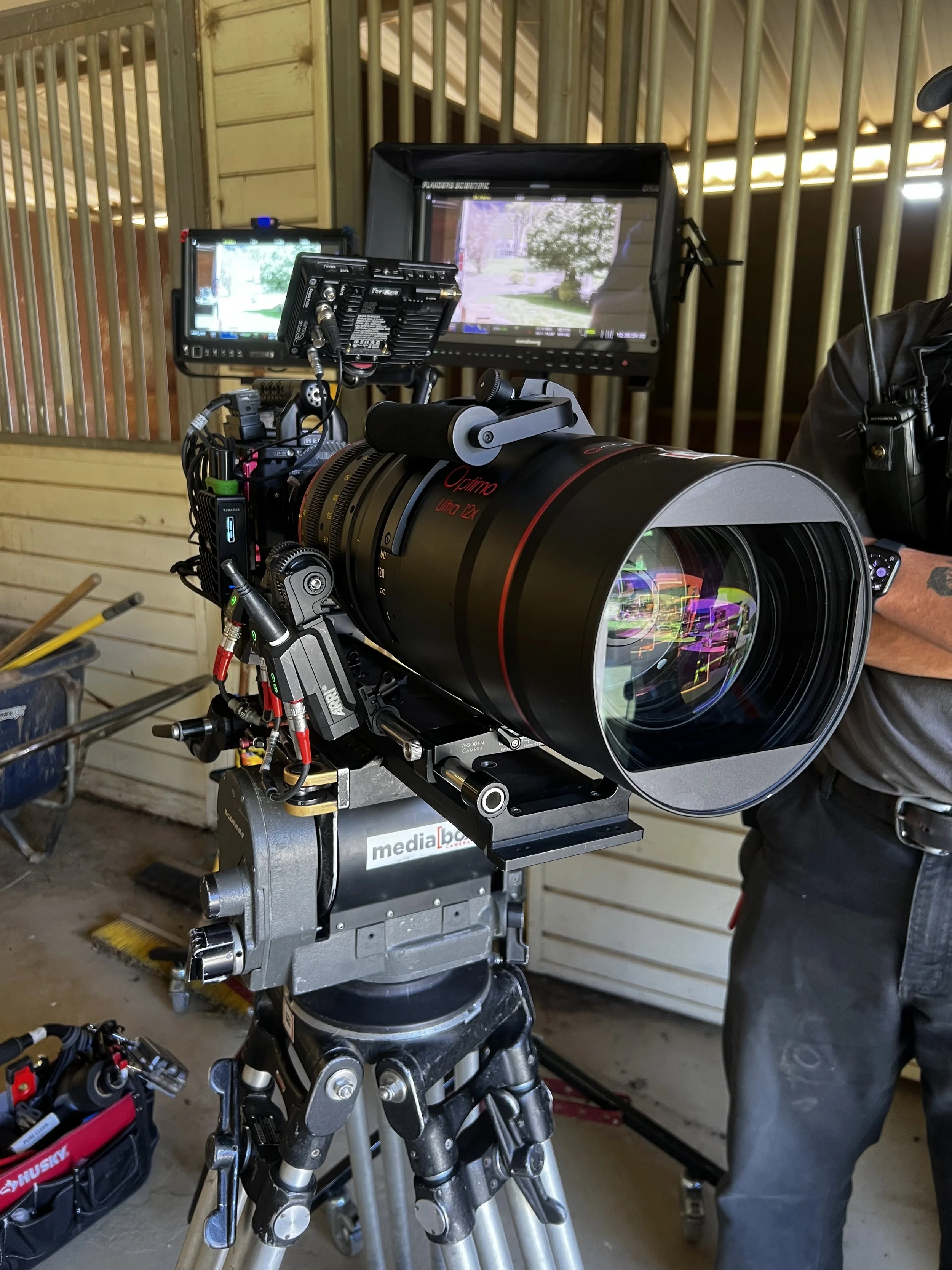 Close-up of a professional camera setup with a large telephoto lens mounted on a tripod, with monitoring screens in the background, in an indoor setting with wooden walls.