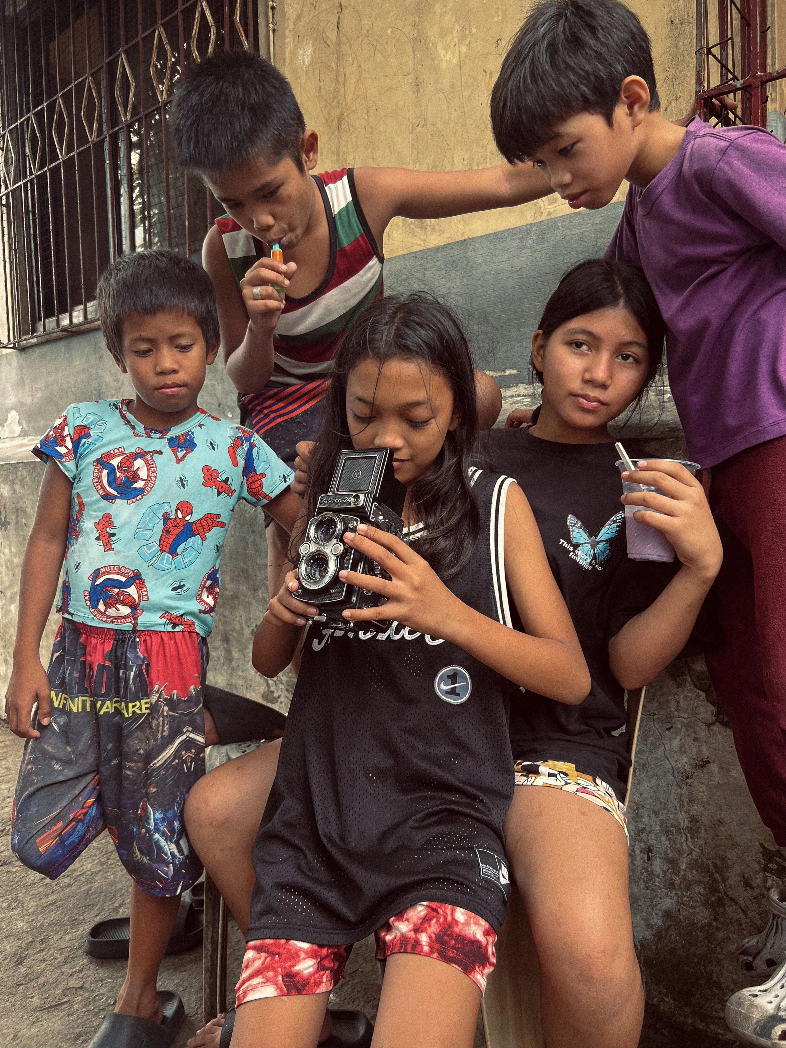 Group of five young children, four boys and one girl, gathered around a girl who is sitting and looking at a camera, while the others look on curiously; they are outdoors near a concrete wall with a fence in the background.