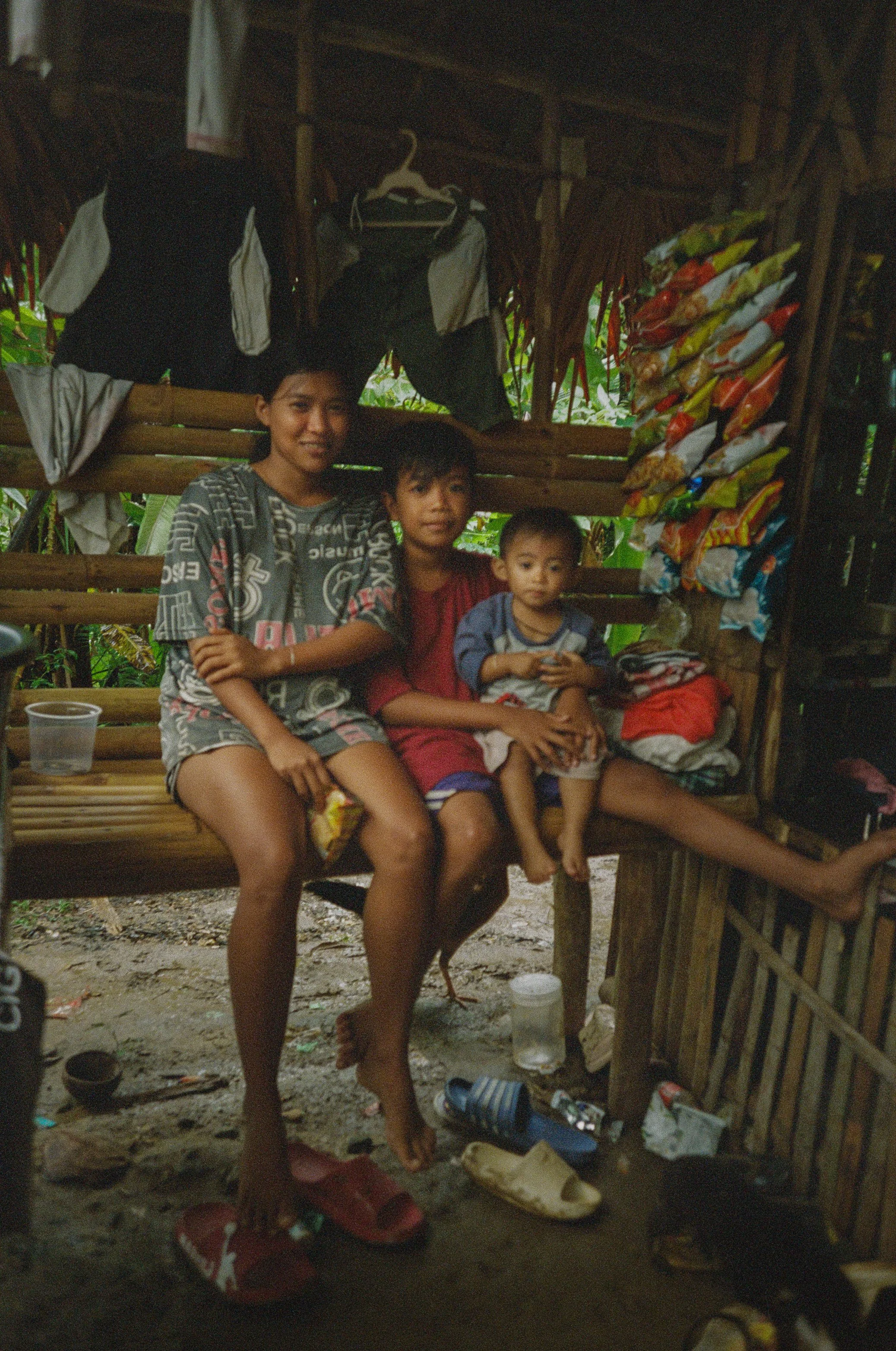 Three children sitting on a wooden bench inside a rustic shelter, with packaging hanging on the wall behind them and various household items on the ground and shelf.