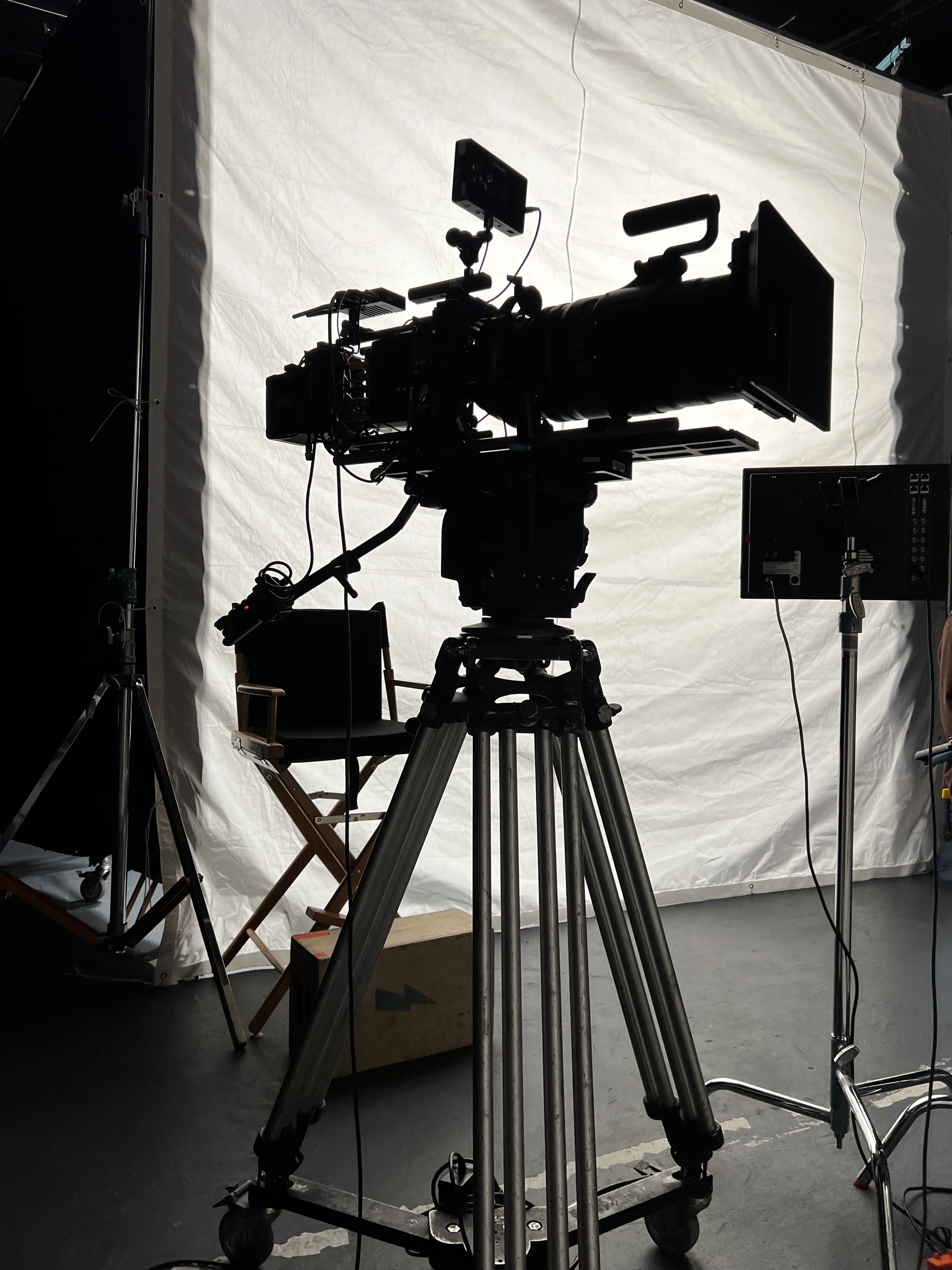 A professional video camera on a tripod with a large lens, facing a white backdrop in a studio or production setting.