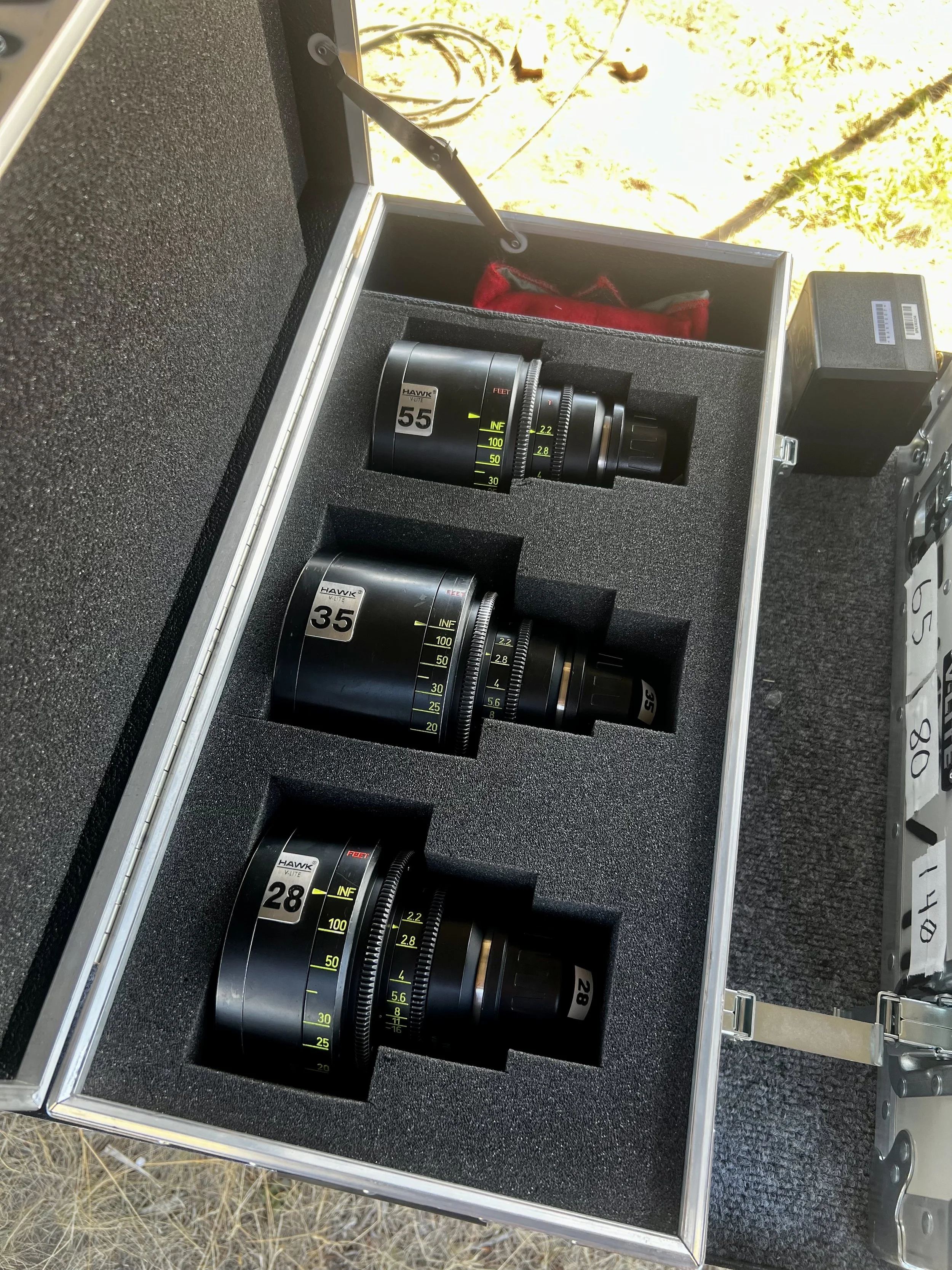 Open carrying case containing three professional camera lenses with numbered labels 55, 35, and 28, resting on a black foam padding. Outside the case, some equipment and grass are visible.